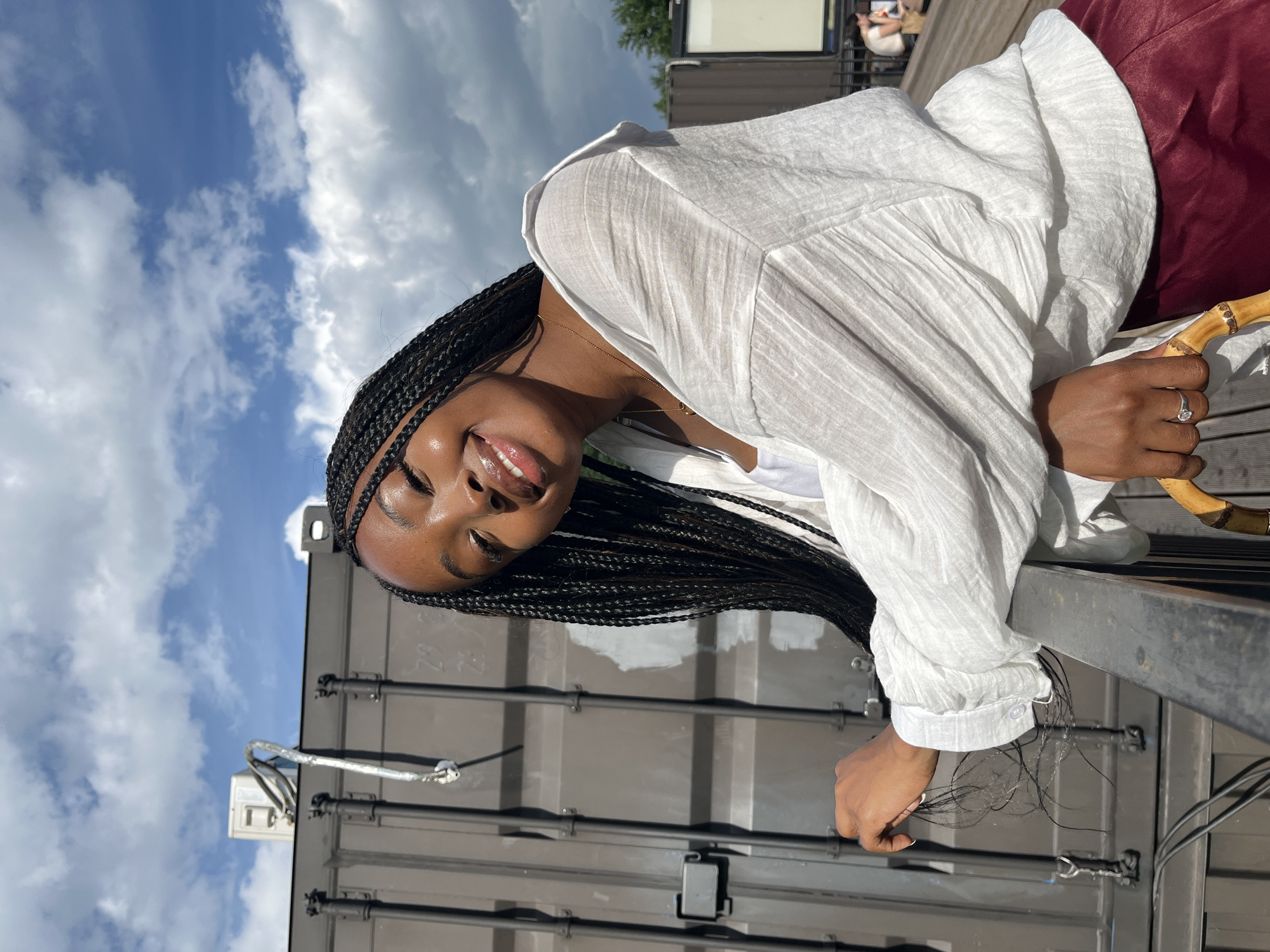 A young Black woman with braided hair leans on a railing, smiling at the camera. She is wearing a white, loose-fitting shirt over a white tank top and a maroon skirt. Her left hand rests on the railing, and her right hand is holding a bamboo-handled object. In the background, a large, dark gray shipping container is visible against a blue sky with white clouds. The scene appears to be outdoors, possibly on a rooftop or elevated platform, with wooden decking in the foreground. There are no discernible landmarks or elements that specifically place this scene in Kazan, Russia.