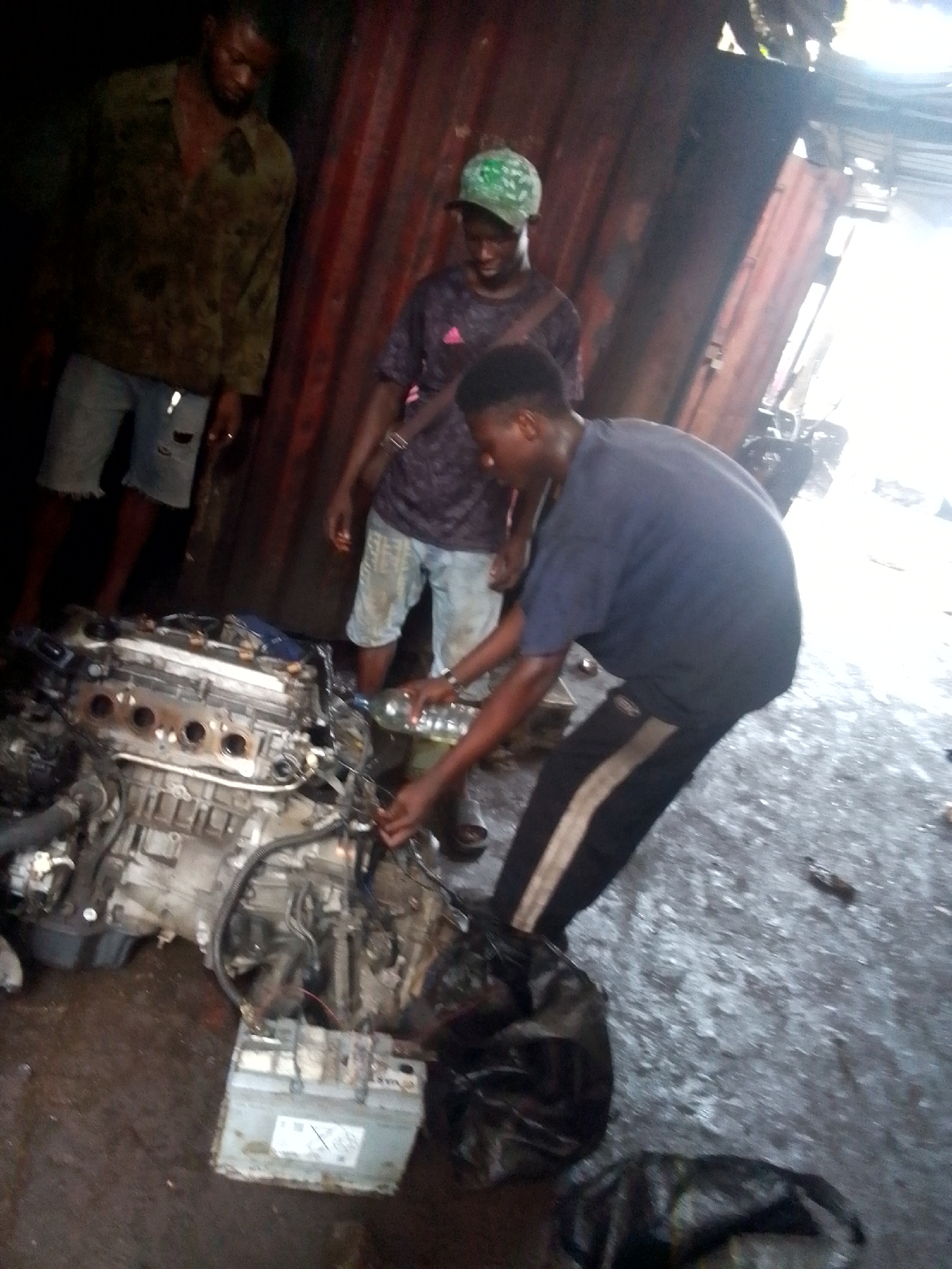 The scene depicts three men gathered around a car engine in a workshop located in Onitsha, Nigeria. The workshop appears to be a semi-enclosed space, with walls of corrugated metal sheeting. The floor is dark and appears to be wet or greasy. To the left, a man is seen wearing a dark olive jacket and jeans. Standing in the center, the second man is wearing a green cap and a blue t-shirt and light-colored jeans. The third man, wearing a blue shirt and black sweatpants, is leaning over the engine, apparently pouring liquid from a bottle onto it. A car battery sits near the engine, and black plastic bags litter the floor. Light streams in from an opening in the wall, creating a bright spot in the background.