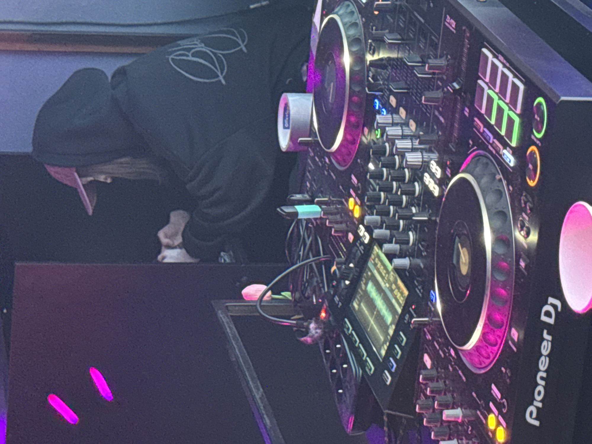 A person in a black hoodie and pink cap is seen bending over a Pioneer DJ system, which is the prominent subject in the image. The DJ system, with its controls and lit jog wheels, is set on a dark surface illuminated by purple light. Parts of a speaker and cables are also visible. The location cannot be determined as being in Komsomolsk-on-Amur, Russia, or any other specific location from the visual information.