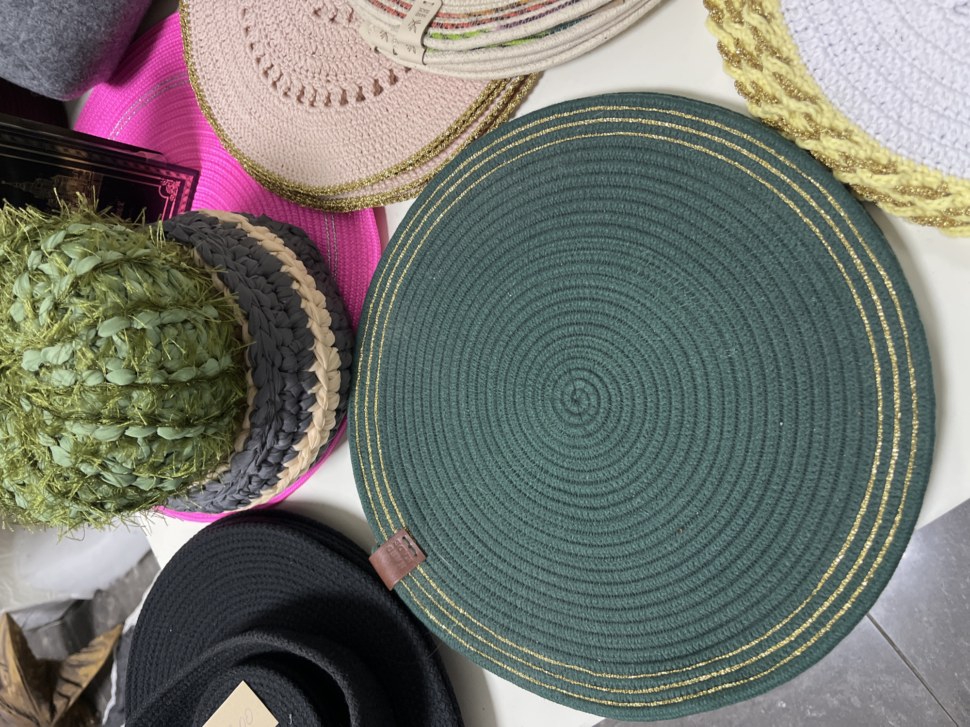 The image displays a collection of woven placemats and a decorative potted plant, likely on a table in a retail setting or home. A prominent dark green, circular placemat with thin gold stripes is in the foreground. To the left, a black woven hat or bowl is partially visible. Behind the green placemat, a pink woven placemat with a textured pattern and gold trim is present. A decorative plant, resembling a cactus, is in a gray and beige woven pot, positioned behind the pink placemat. Other colorful woven placemats are visible towards the right and bottom of the frame. The overall setting appears to be indoors, with a neutral background. There are no people or actions depicted. The items are arranged for display. The image does not contain any identifiable landmarks or clues to indicate its specific location within the city of Hrodna, Belarus.