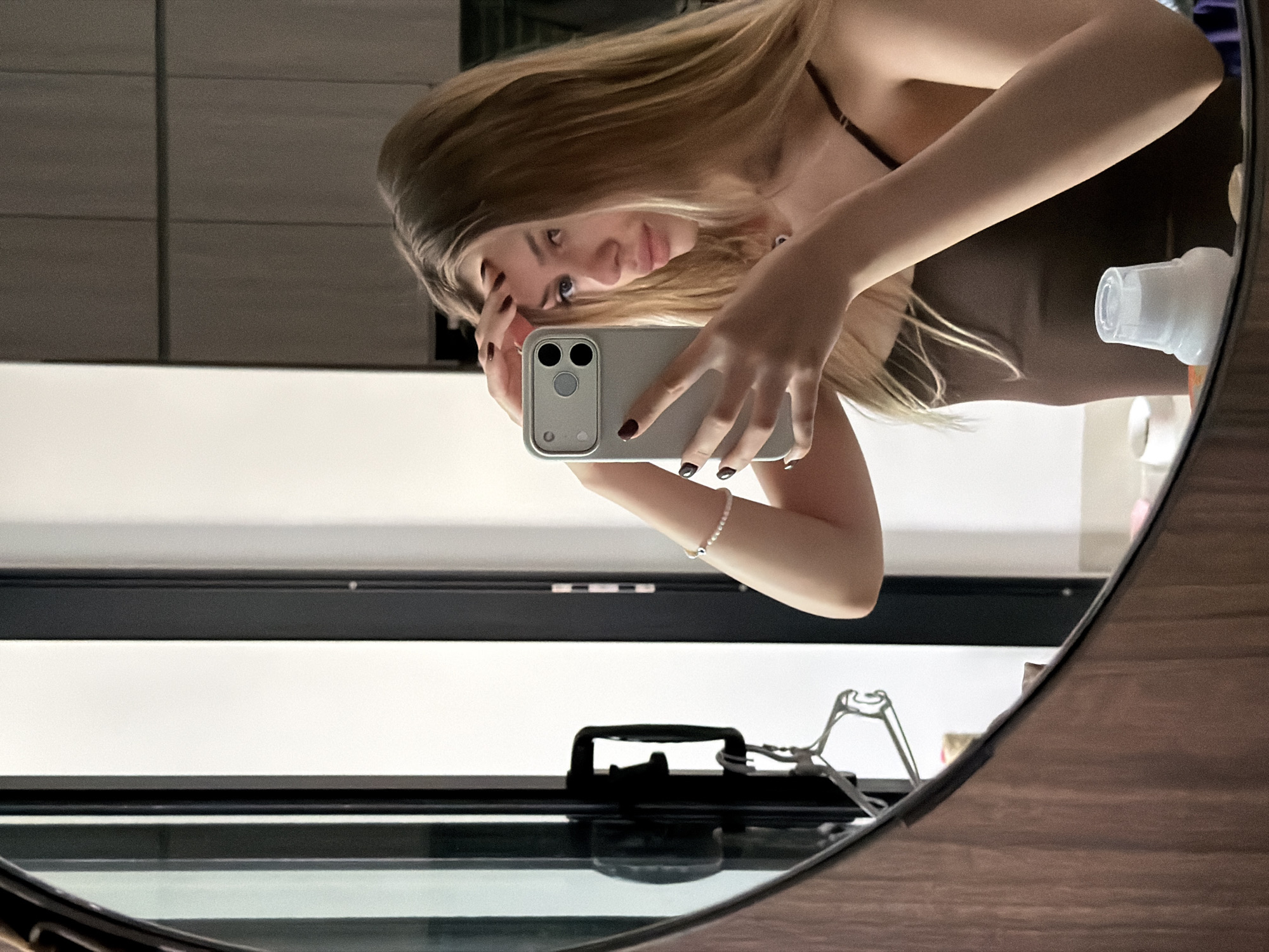 A young woman with long blonde hair is taking a mirror selfie in what appears to be a modern bathroom or dressing area. She is wearing a dark brown spaghetti strap top and has dark red nail polish. Her left hand is holding her phone, a silver iPhone with a light-colored case, while her right hand is touching her forehead, possibly adjusting her hair. Her gaze is directed at the phone's camera. In the reflection of the round, wooden-framed mirror, we can see a tall, dark framed doorway or closet with a white interior. There's a black handle on the door and a clothes hanger is attached to it. Further back, a minimalist kitchen or vanity area with dark cabinetry is visible. A white plastic bottle with a translucent cap is also in the foreground, reflected in the mirror. The overall lighting is soft and diffused, creating a relaxed ambiance. The image does not contain any information that can place the location in Đà Nẵng, Vietnam.