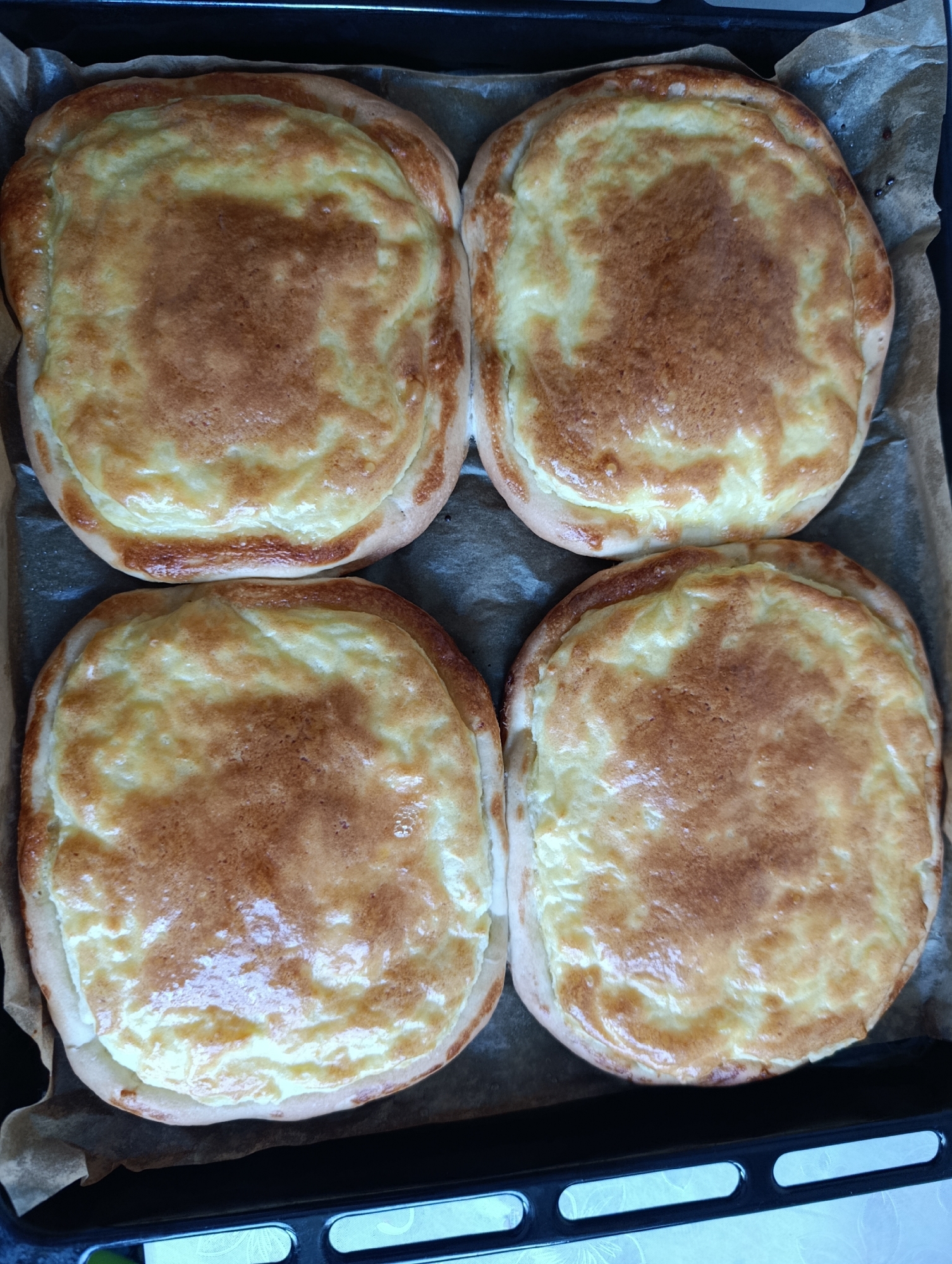 Four cheese-filled pastries are arranged on a baking sheet lined with parchment paper, inside a dark baking tray. The pastries have a golden-brown crust and a creamy, slightly browned filling. They are viewed from directly above. The location is unknown.