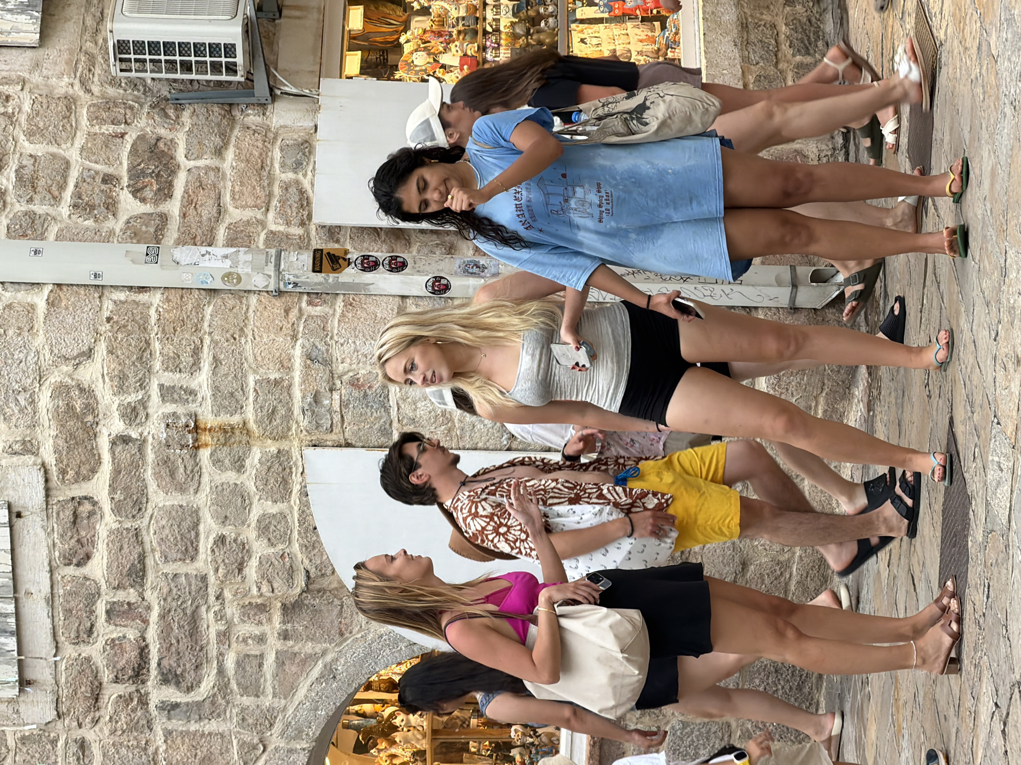 In Budva, Montenegro, a group of young adults walks on a cobblestone street in front of a stone building. A woman in a pink top and black shorts carries a cream-colored tote bag. Next to her, a man in a floral shirt and yellow shorts walks with his hands at his sides. Another woman in a grey tank top and black shorts holds a phone. To her right, a woman in a light blue oversized t-shirt walks with a patterned tote bag and is touching her face. Further back, a woman in a dark top and skirt is visible, along with other figures in the background. The building is constructed of rough stone blocks, and an air conditioning unit is mounted on the exterior wall. Various shops with visible merchandise are located in the background.