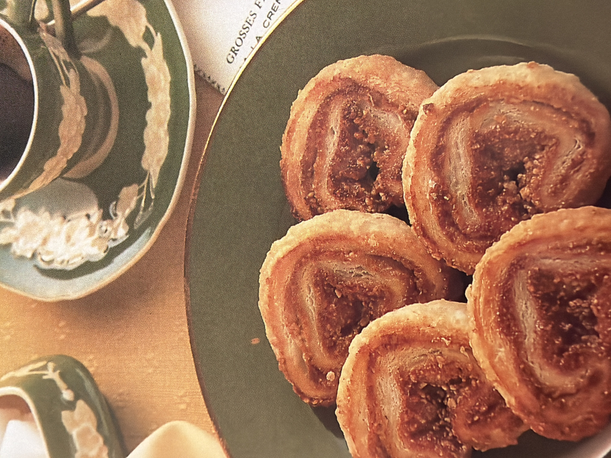 A still life photograph showcases a close-up view of baked pastries on a dark green plate. The pastries are circular and appear to be spiral-shaped, with layers of flaky dough and a filling visible in the center. Four of these pastries are arranged on the plate, with one in the foreground partially obscuring the others.
In the upper left quadrant of the image, a small, green ceramic dish with a decorative pattern holds several white sugar cubes. To the right of this dish, a green ceramic teacup with a saucer, also decorated with a floral design, holds a dark liquid, likely coffee or tea. A portion of a printed card or menu is visible in the lower right corner, with the words "GROSSES F" and "A CREM" partially legible.
The background is a textured, golden-orange surface, suggesting a tablecloth or placemat. The lighting is warm and casts subtle shadows. The overall scene evokes a sense of a delicate breakfast or afternoon tea setting. The image is sharp and detailed, with a shallow depth of field that blurs the background and highlights the pastries.