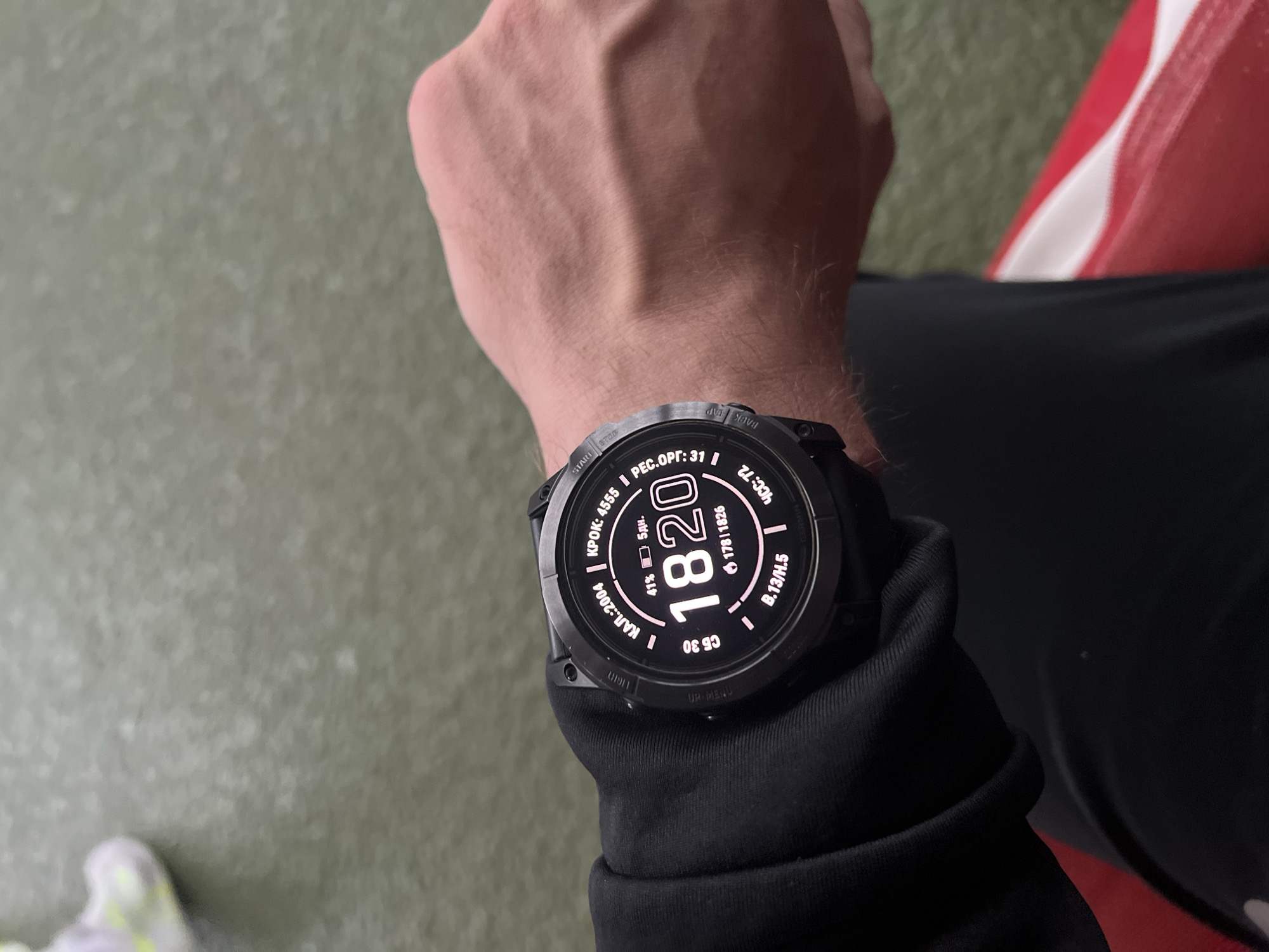 A person's left wrist is adorned with a black smartwatch displaying "18:20" (6:20 PM) as the time. The watch face also shows indicators for battery level (41%), steps (4555), calories (2004), heart rate (72 bpm), and other fitness metrics. The person is wearing a black long-sleeved garment. The background includes a surface, possibly flooring or ground, with a greenish hue, and a red and white object is visible on the upper right side. No specific information indicates this scene is located in the city of Poltava, Ukraine.