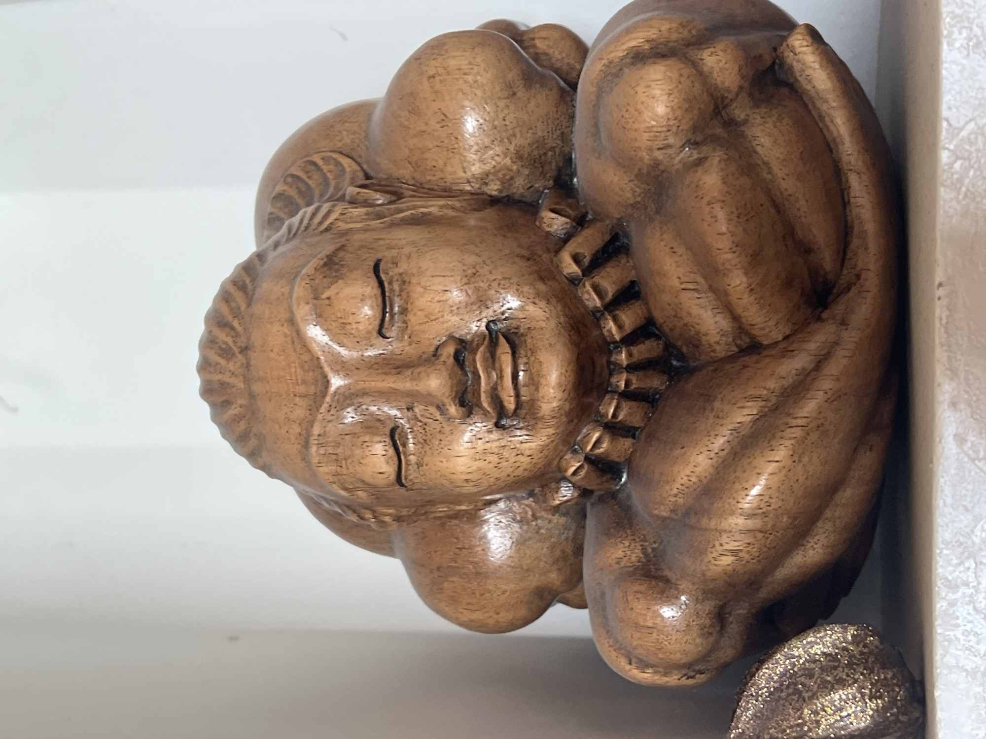 This is a close-up, vertical shot of a carved wooden figurine of a person in a fetal position, with their arms wrapped around their head and body. The wood is a warm, brown tone with visible grain patterns and a glossy finish. The figurine appears to be sleeping or resting, with closed eyes and a relaxed expression on its face. The carving details are intricate, particularly around the head and shoulders, where there are textured areas resembling hair or fabric. The background is a plain white surface, suggesting the object might be displayed on a shelf or table. There is also a glimpse of another object in the lower right corner, which is dark and appears to be textured or glittered. The lighting is soft and diffused, creating subtle highlights and shadows on the wood. No text is visible in the image.