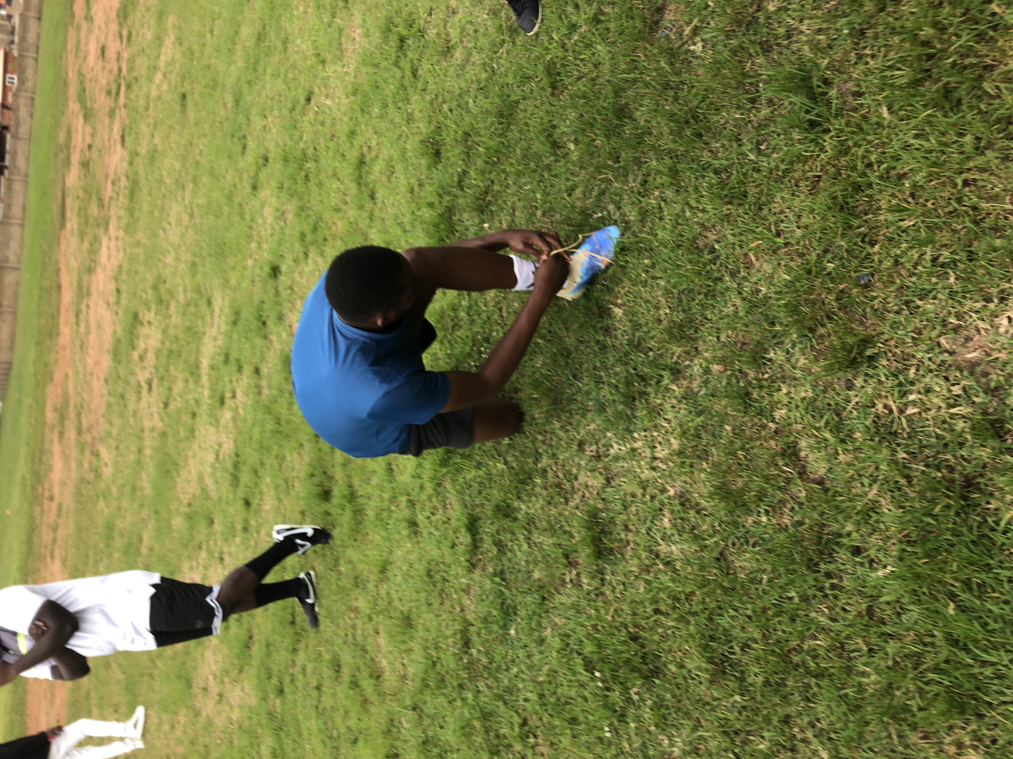 A young Black male wearing a blue t-shirt and black shorts kneels on a grassy field, tying the laces of a colorful soccer cleat on his right foot. The cleat has blue, yellow, and white accents. He is wearing white socks. In the background, another Black male stands with his arms crossed, wearing a white t-shirt, black shorts with white trim, black socks, and black soccer cleats. A third individual, partially visible on the far left, wears white pants and white sneakers. The environment appears to be an outdoor sports field with dry, reddish-brown patches of earth visible amongst the green grass. In the distance, a brick building with a corrugated metal roof is visible. The scene takes place in Jos, Nigeria, possibly at a sports training facility or park.