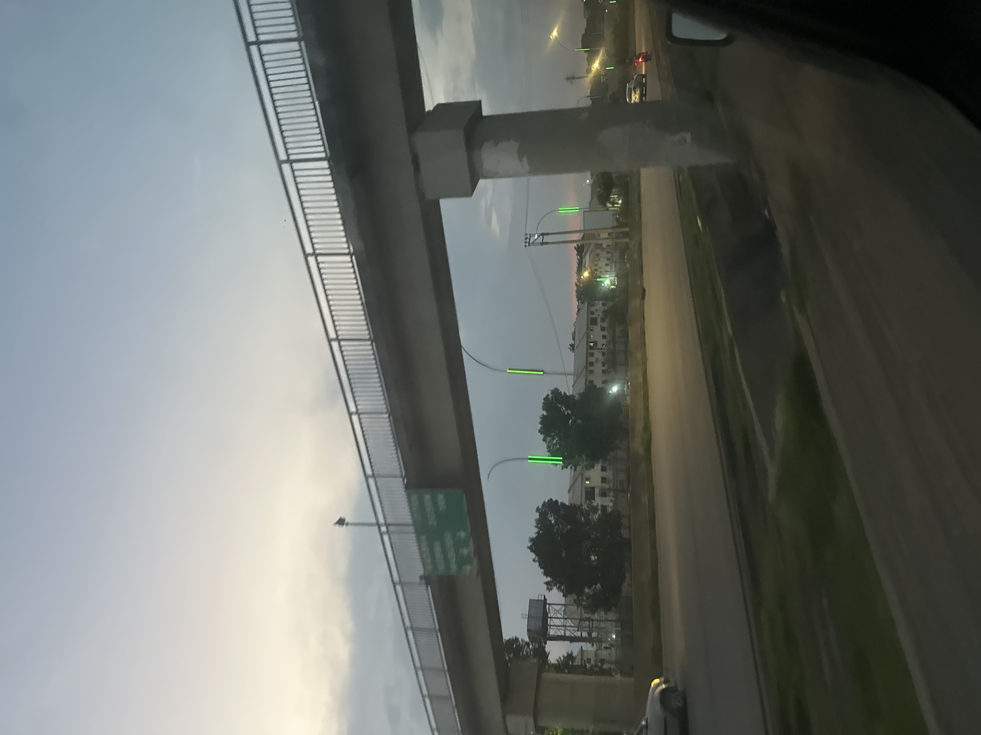 The image shows an overpass in Ade, Nigeria, viewed from a car. The overpass is made of concrete and has a railing on the side. There are streetlights with green lights on the side of the road. In the background, there are buildings and trees. The sky is cloudy, and it appears to be dusk. There are cars on the road. A green sign with white text is visible on the underside of the overpass, but the text is not legible. The overall impression is that of a typical urban scene in Nigeria during twilight.