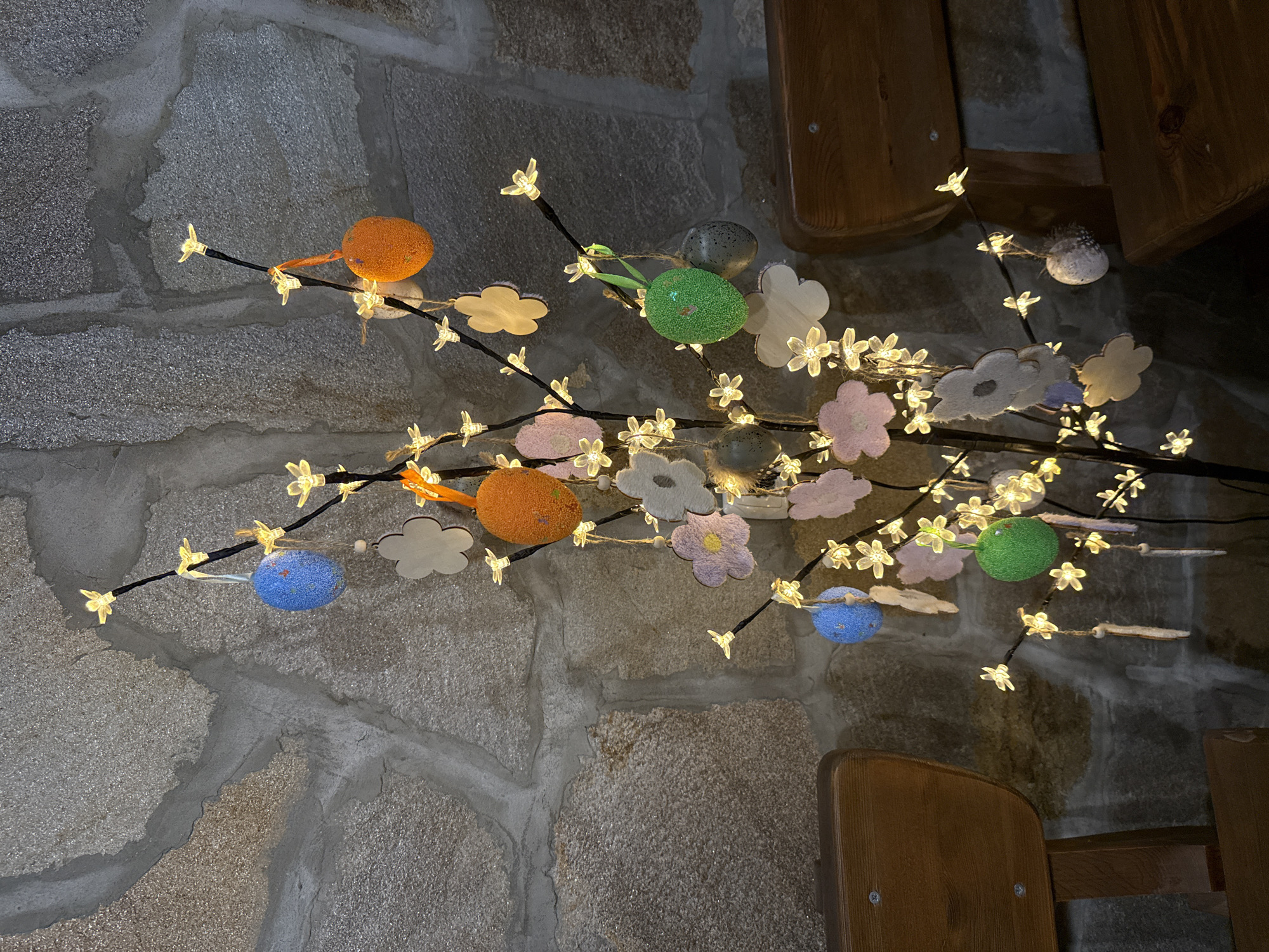 The image shows an illuminated decorative tree with colorful Easter egg ornaments and flower-shaped decorations, likely indoors against a stone wall. Parts of wooden furniture are visible in the lower right of the frame. The location is in the city of Lukovit, Bulgaria.