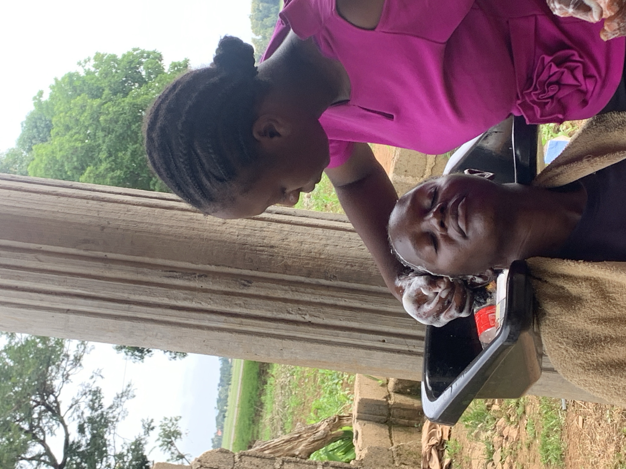 In Agban, Nigeria, two women are depicted in an outdoor setting. The woman on the right, wearing a bright pink off-the-shoulder top with floral embellishments, is tending to the woman on the left. The woman on the left, with her eyes closed and a towel draped around her neck, is seated with her head tilted back over a black basin, suggesting she is having her hair washed. The woman on the right is seen with her hand, covered in soap suds, gently washing the hair of the woman in the basin. In the basin, a red and white bottle is visible. The background shows a large, fluted concrete pillar, greenery, and a distant rural landscape with fields and scattered buildings. The overall scene appears to be a makeshift salon or a residential area where hair services are being provided.
