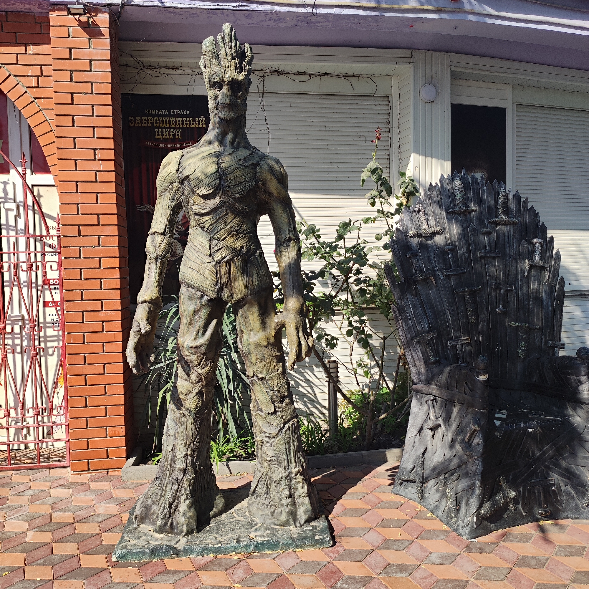 The image shows a statue of Groot, a character from Marvel's Guardians of the Galaxy, standing next to a replica of the Iron Throne from the TV series Game of Thrones. Both are placed outdoors on a brick-patterned walkway. Behind the statue and throne is a building with a white facade and a sign that reads "КОМНАТА СТРАХА" (House of Horrors) and "ЗАБРОШЕННЫЙ ЦИРК" (Abandoned Circus). The scene appears to be an attraction or photo opportunity. The lighting suggests it is daytime. There are no people visible in the image.