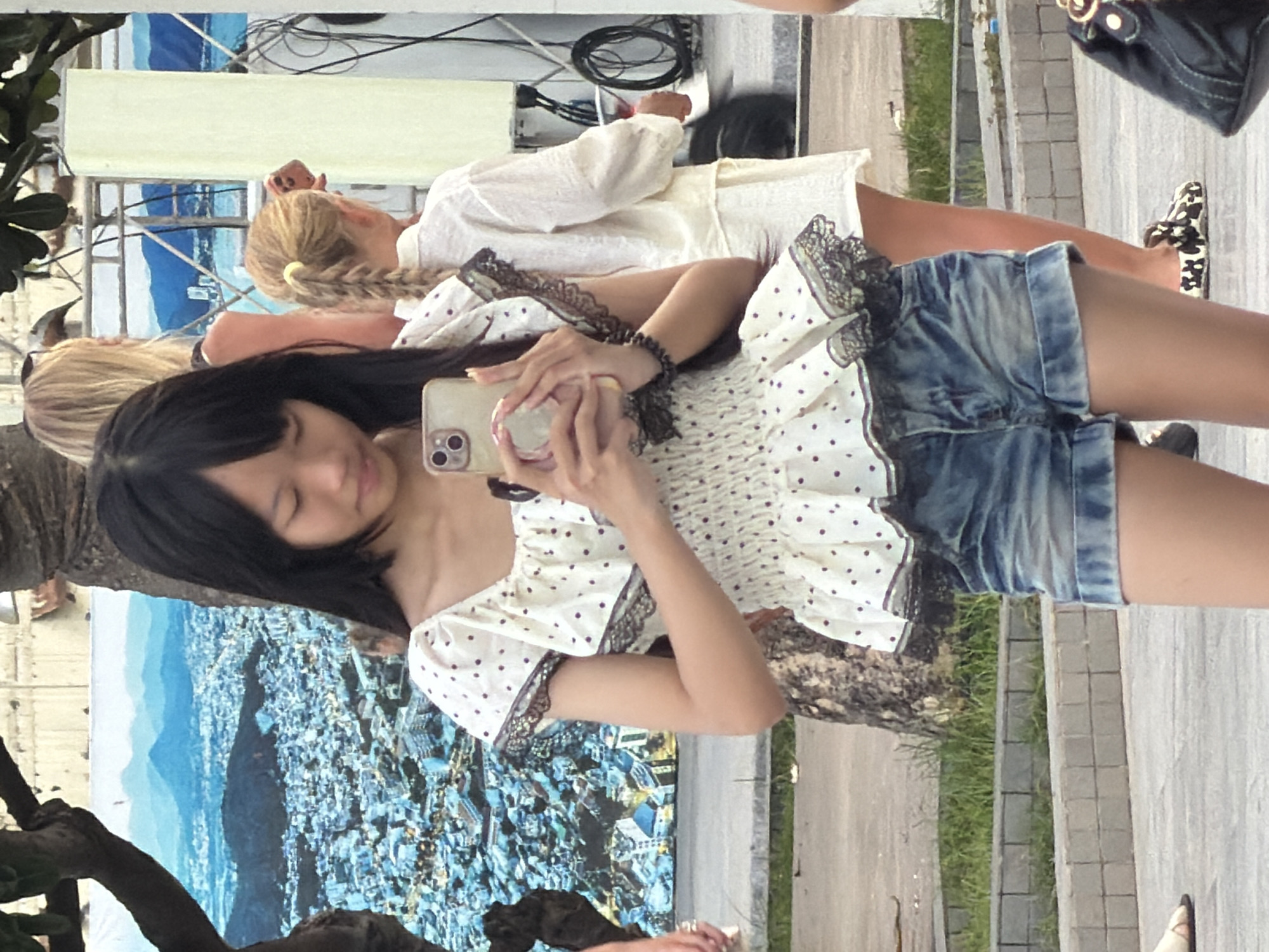 A young Asian female with long dark hair is holding a smartphone and taking a selfie. She is wearing a white off-the-shoulder blouse with purple polka dots and ruffled trim, and distressed denim shorts. She is standing on a paved area with a grassy strip and a tiled border behind her. In the background, two other females are visible. One, with blonde hair in a braid, wears a white shirt and white shorts. Another, with blonde hair, wears a light-colored top. A large mural of a cityscape and mountains is visible behind the primary subject. The scene appears to be in an outdoor public space, possibly near a waterfront or promenade, consistent with locations in Nha Trang, Vietnam, given the tropical vegetation and mural style.