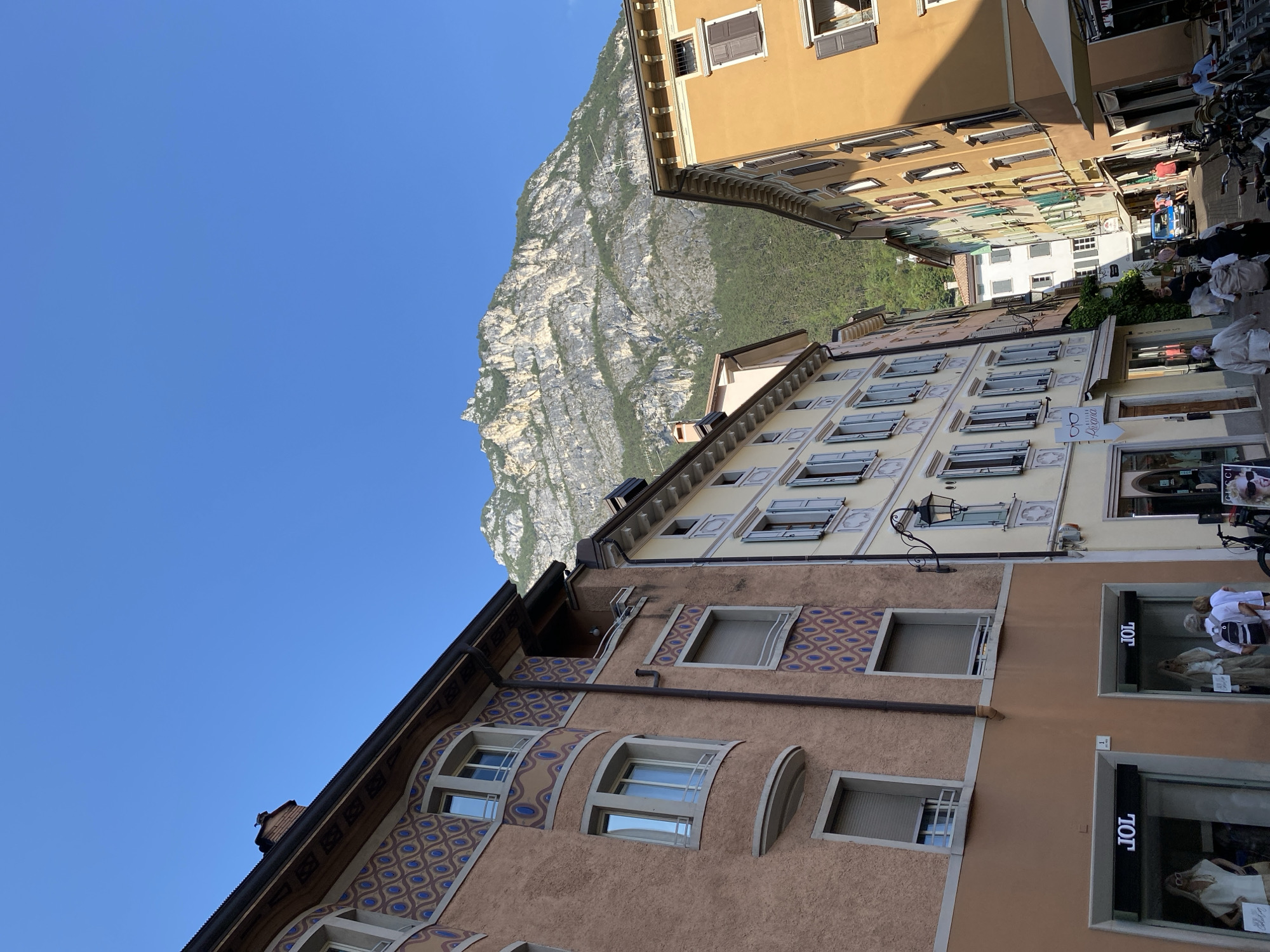 A low-angle, eye-level shot captures a street scene in Riva del Garda, Italy, under a clear blue sky. The foreground is dominated by the facades of several buildings. On the left, a building with a reddish-brown textured facade features decorative blue and brown patterns around its arched windows. Below, two storefronts, both labeled "JOL," are visible. To the right of this building, a lighter-colored building with cream-colored trim and light blue shutters stands. Further down the street, the buildings continue in a variety of colors, including yellow and orange.

In the background, a large, rugged mountain rises steeply, covered in dense green trees at its base and showing rocky, gray slopes higher up. The mountain dominates the upper portion of the image.

Several people are present in the scene. Two figures are walking away from the camera on the sidewalk in front of the "JOL" stores. Further down the street, other pedestrians can be seen walking. A blue car is visible in the distance, navigating the street. Bicycles are parked along the side of the street. An awning shades an outdoor seating area on the right side of the frame. The street itself appears to be paved.