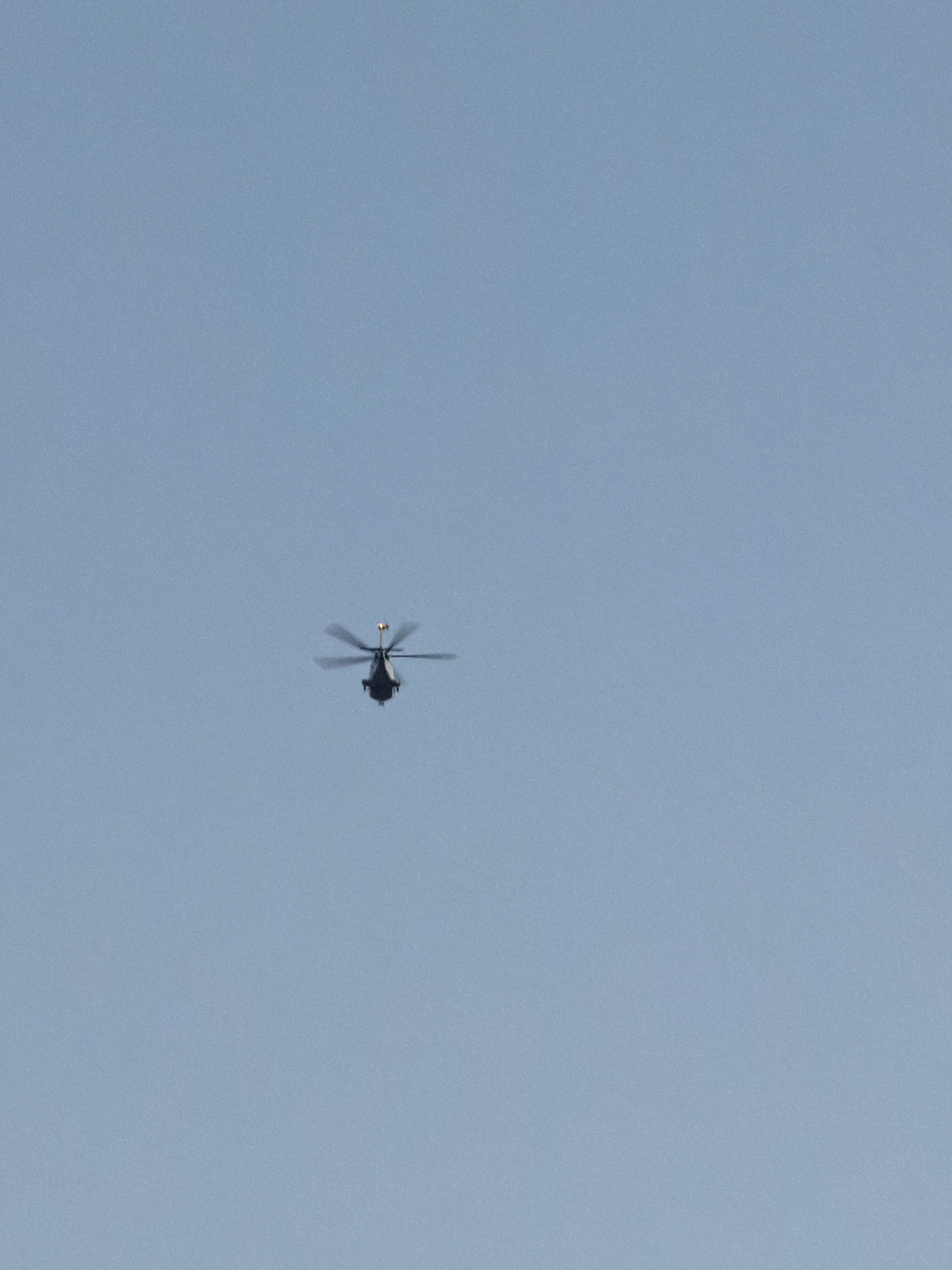 A dark-colored helicopter with its rotors in motion is captured in flight against a clear, pale blue sky. The helicopter appears to be at a significant altitude and is positioned in the upper right quadrant of the image. The lighting suggests it is daytime. The scene does not contain any identifiable landmarks or elements that would place it specifically in the city of Salou, Spain.
