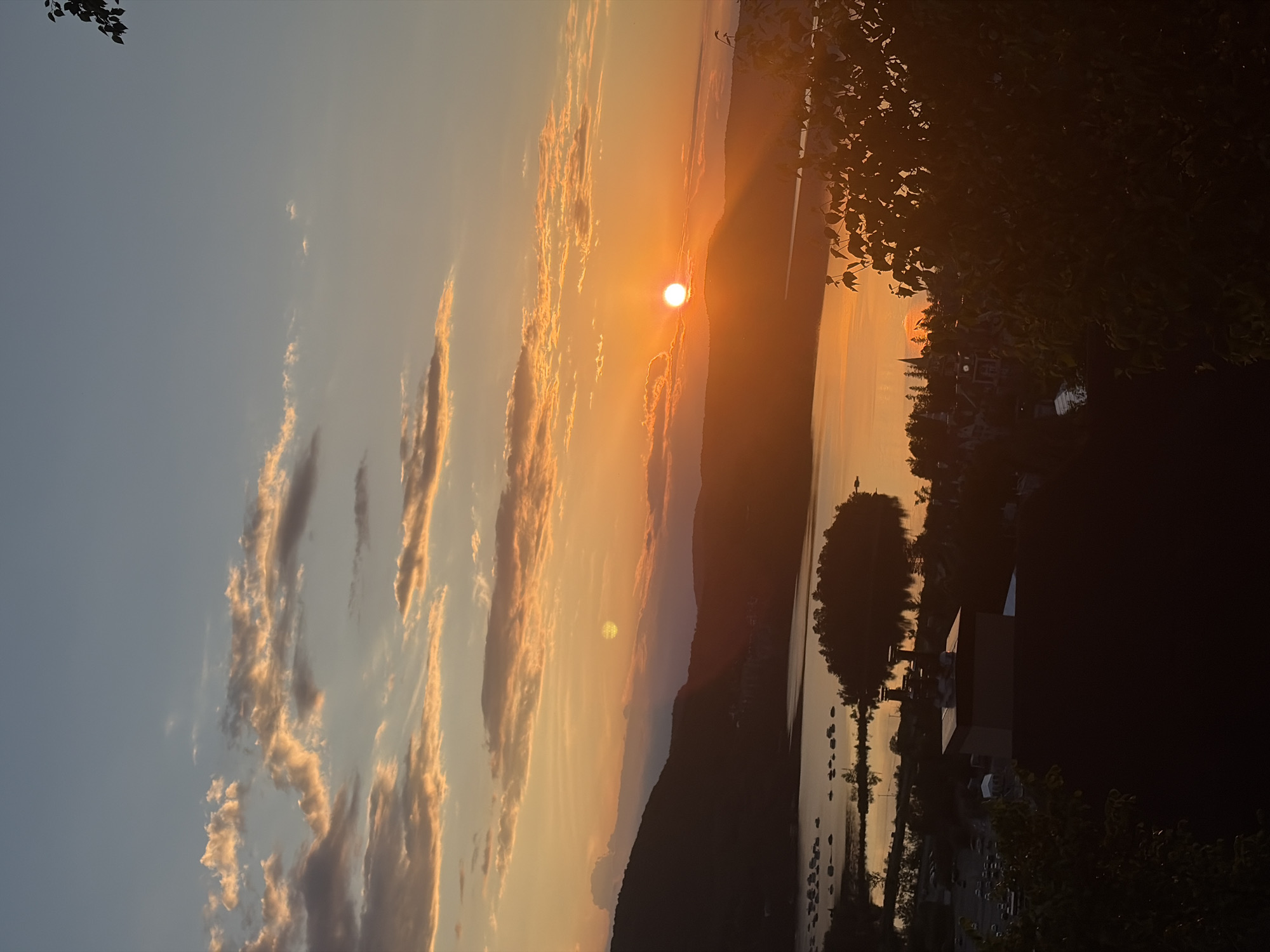 This is a vertical, eye-level, outdoor, full shot photograph of a sunset over a body of water, likely Lac Tremblant, in Mont-Tremblant, Canada. The sun is a bright, orange orb positioned just above the horizon, casting a warm glow over the landscape. Wispy, golden-orange clouds are scattered across the sky, contrasting with the clear blue sky above. Silhouetted mountains form the backdrop, their dark forms softened by the evening light. In the foreground, a body of water reflects the sunset's colors, with a small island dotted with trees in the middle. On the shore of the lake, there are visible boats, as well as buildings and a parking lot. The image is taken from an elevated vantage point, looking down on the scene, with dark trees in the lower right corner framing the view. The overall impression is one of tranquility and natural beauty.