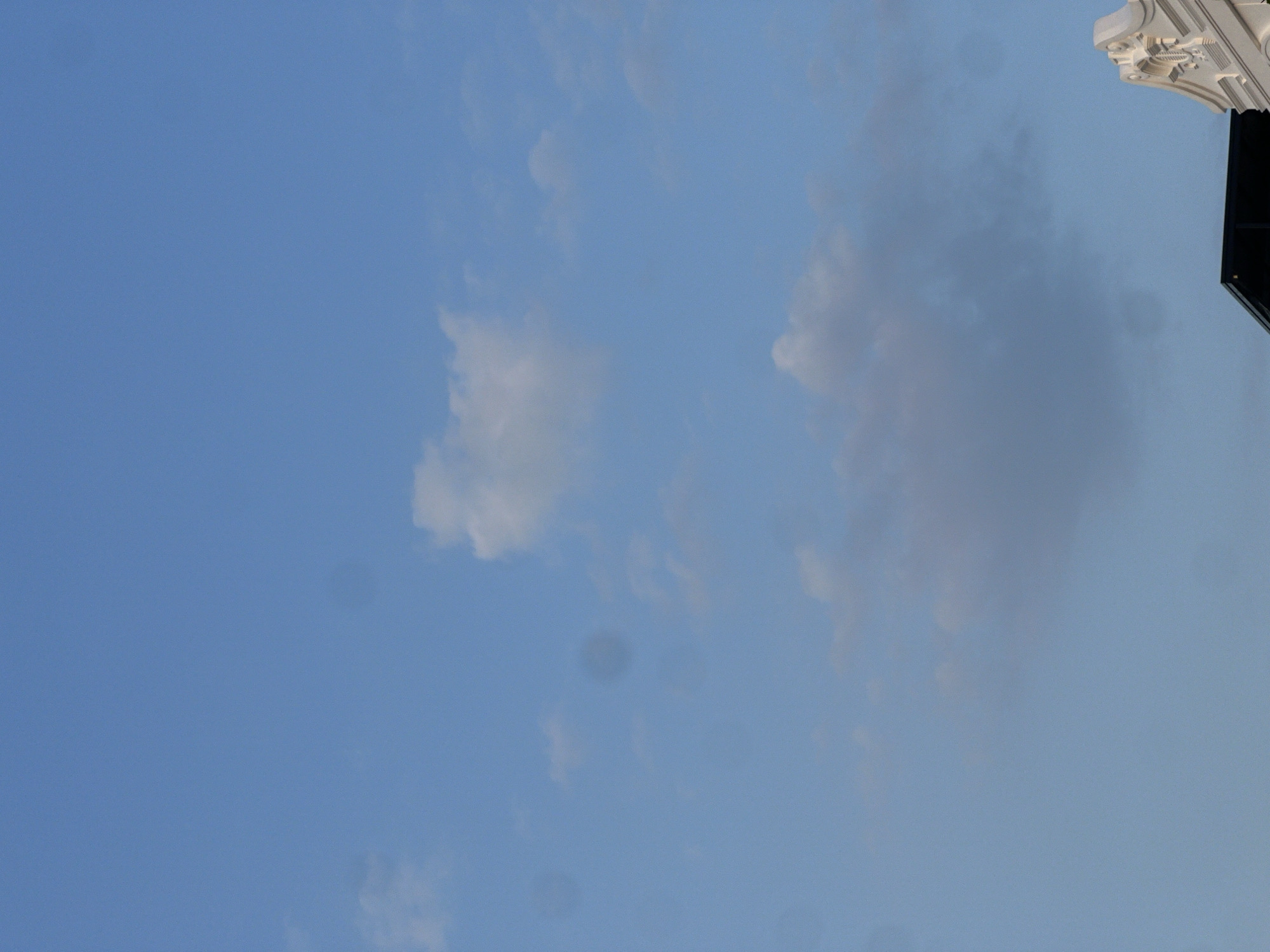 The image shows a view of the sky in Nam Nha Trang, Vietnam. The sky is a pale blue with scattered clouds. A section of a white building with decorative architectural details is visible in the upper right corner of the image. The light suggests it is either early morning or late afternoon. The image is slightly blurred, indicating it may have been taken through a window.