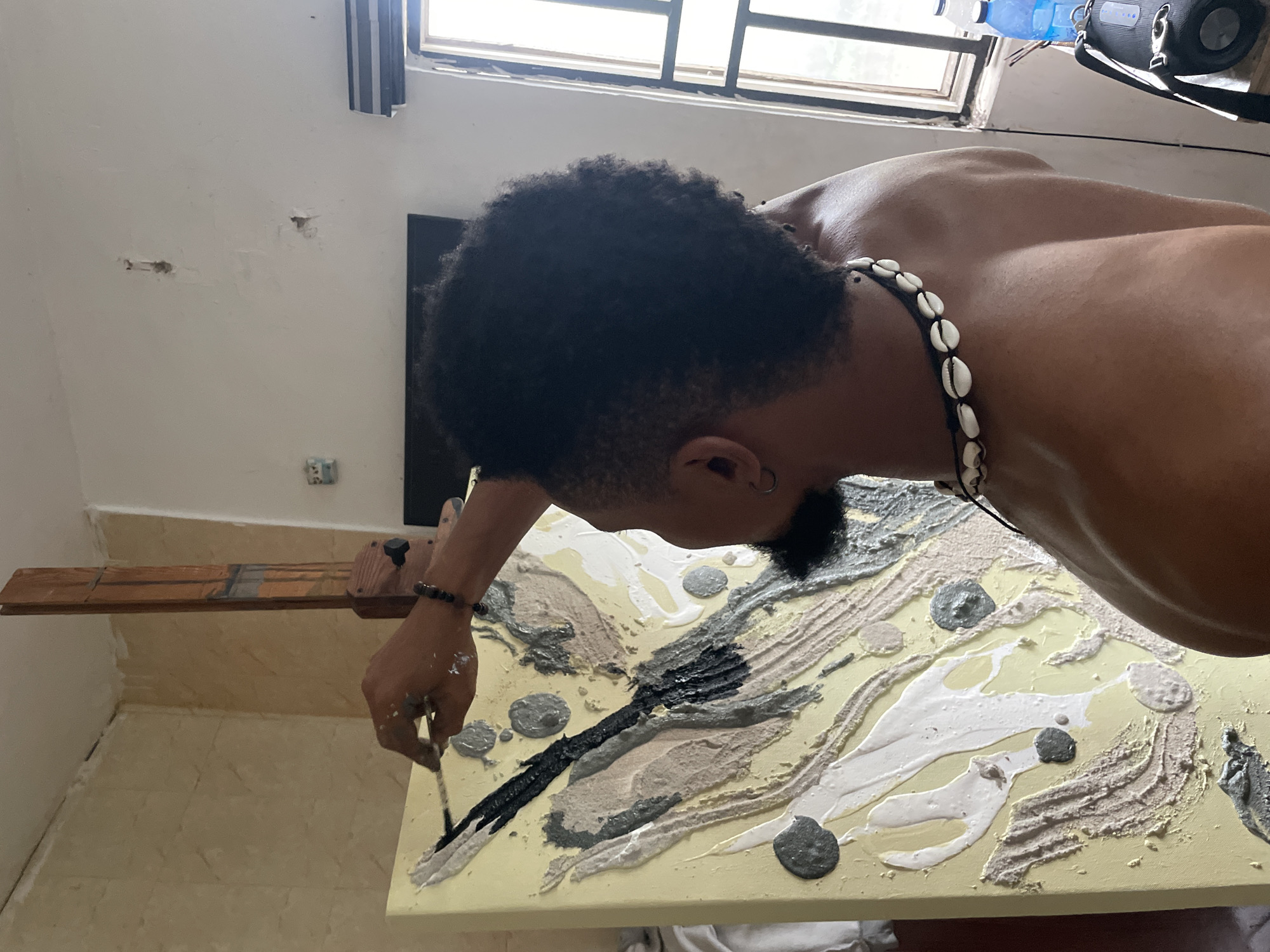 The image shows a person painting on a canvas. The person has dark hair, a goatee, and is wearing a necklace made of shells. The painting has a yellow background with textured strokes of white, grey, black, and tan paint. The person is holding a thin brush and appears to be adding black paint to the canvas. The painting is resting on a wooden structure. The environment appears to be indoors, possibly a studio, with beige walls and tiled floors. A window is visible in the background. Location: Ado, Nigeria.