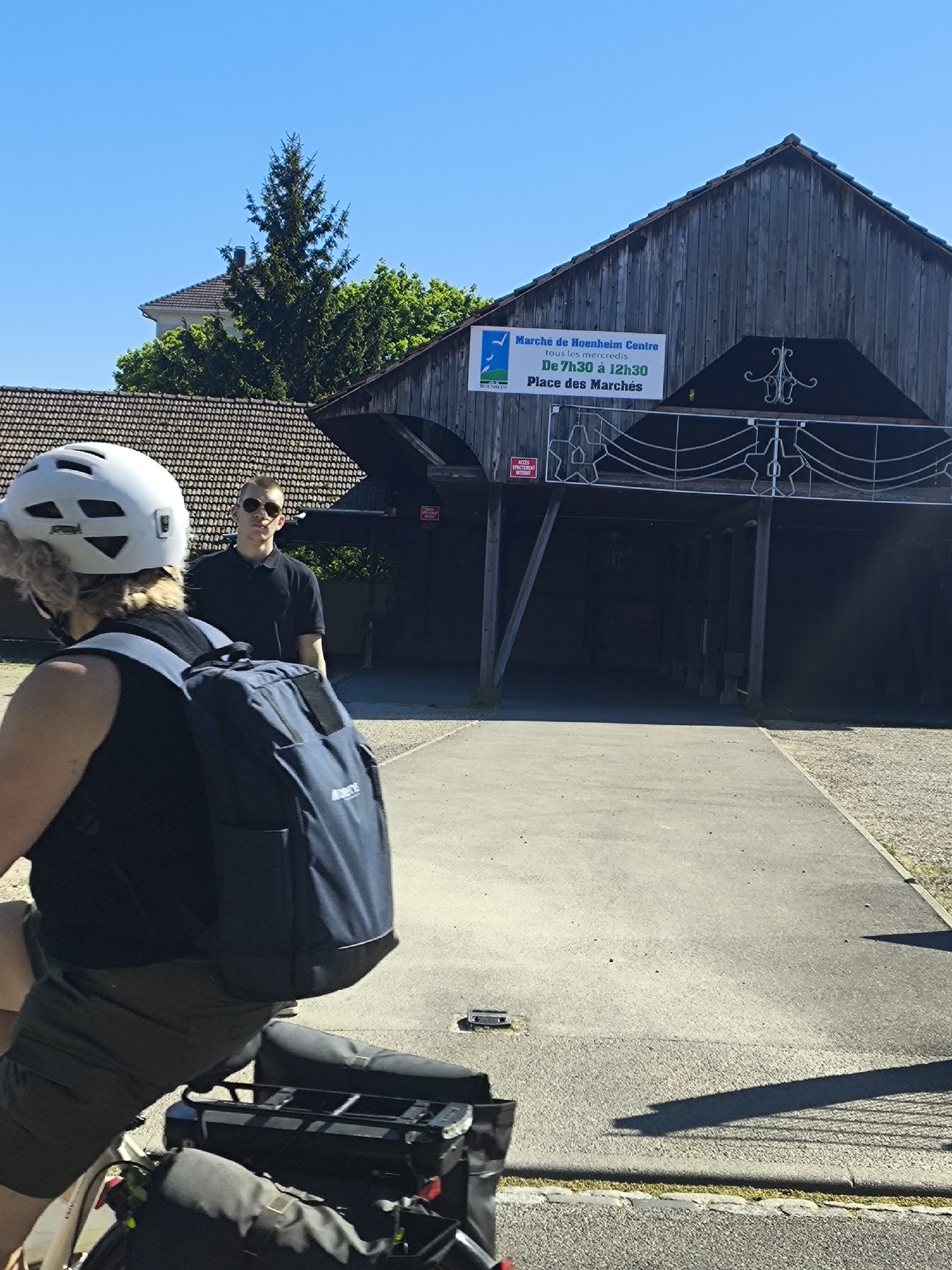 The image depicts a scene in Hoenheim, France, in front of the "Marché de Hoenheim Centre." A woman on a bicycle, wearing a white helmet and carrying a blue backpack, is in the foreground. Standing behind her and to the side of a building is a man in a black shirt and sunglasses. The building has a sign that reads "Marché de Hoenheim Centre, tous les mercredis, De 7h30 à 12h30, Place des Marchés." The building's architectural style is traditional, possibly wooden, with a steeply pitched roof. The foreground consists of a paved pathway. In the background, there is greenery including a tall conifer tree and building with a brown tile roof. The day is sunny.