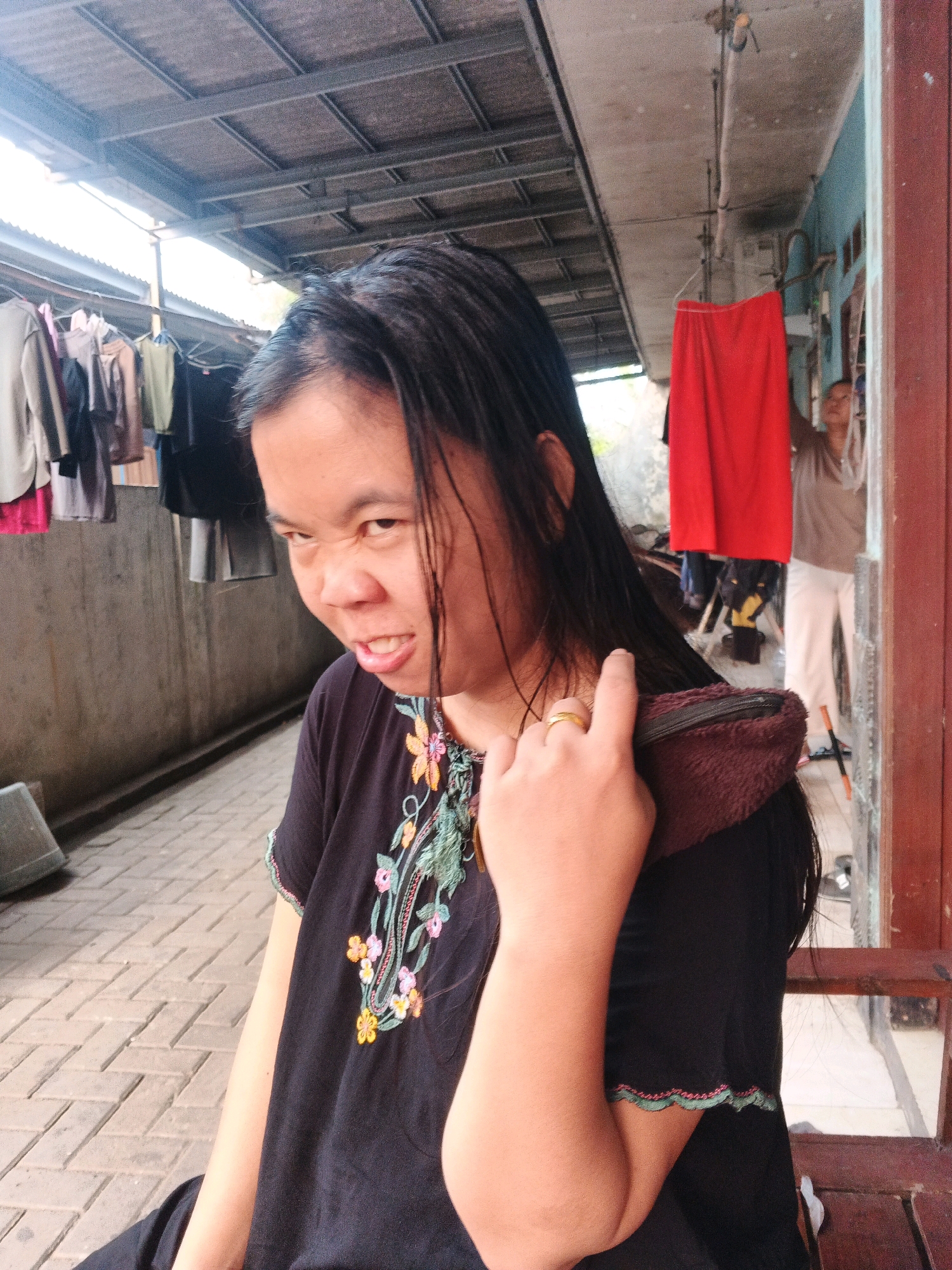 A medium shot shows a woman with dark hair and a black t-shirt with colorful embroidery, sitting outdoors and making a face at the camera. She has a ring on her left hand, which she is holding up near her face and touching her hair. Behind her, clothes are hanging on a line to dry under a corrugated metal roof. A person wearing white pants and a brown shirt is visible in the background. The ground is paved with interlocking bricks, and there is a concrete wall on the left side of the image. The setting appears to be a residential backyard or alley in Kadu, Indonesia.