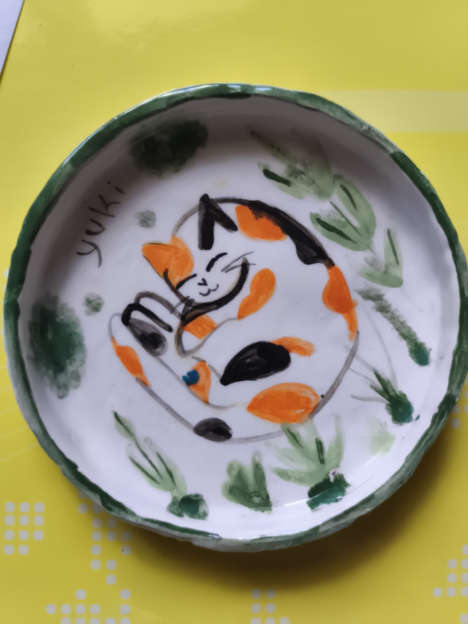A handmade ceramic plate with a green rim is displayed on a yellow surface. The plate features a painted calico cat curled up in the center. The cat is depicted with black, orange, and white patches, with black outlines and facial features. Green watercolor-like strokes, representing foliage, are painted around the cat. The word "Yuki" is written in black script on the upper left side of the plate. The yellow surface has a pattern of small, evenly spaced white dots. This object is not located in the city of Saint-Denis, France.