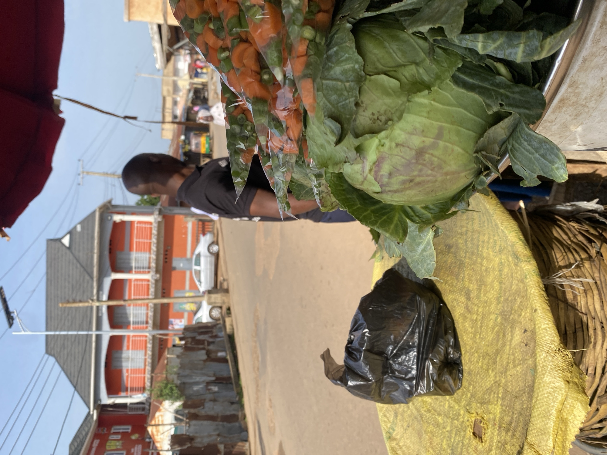 A street vendor in Jos, Nigeria displays fresh produce under a red awning. In the foreground, a large cabbage sits in a metallic container, its green leaves partially obscuring the top of the container. To its left, a black plastic bag lies on a yellow fabric covering a basket. Behind the produce, a man in a black t-shirt is partially visible, facing away from the camera. In the background, a street runs through the city, with buildings and a few vehicles, including a silver car, lining its sides. A solar-powered street lamp stands tall on the side of the road. The overall scene depicts a bustling market atmosphere in an urban Nigerian setting.