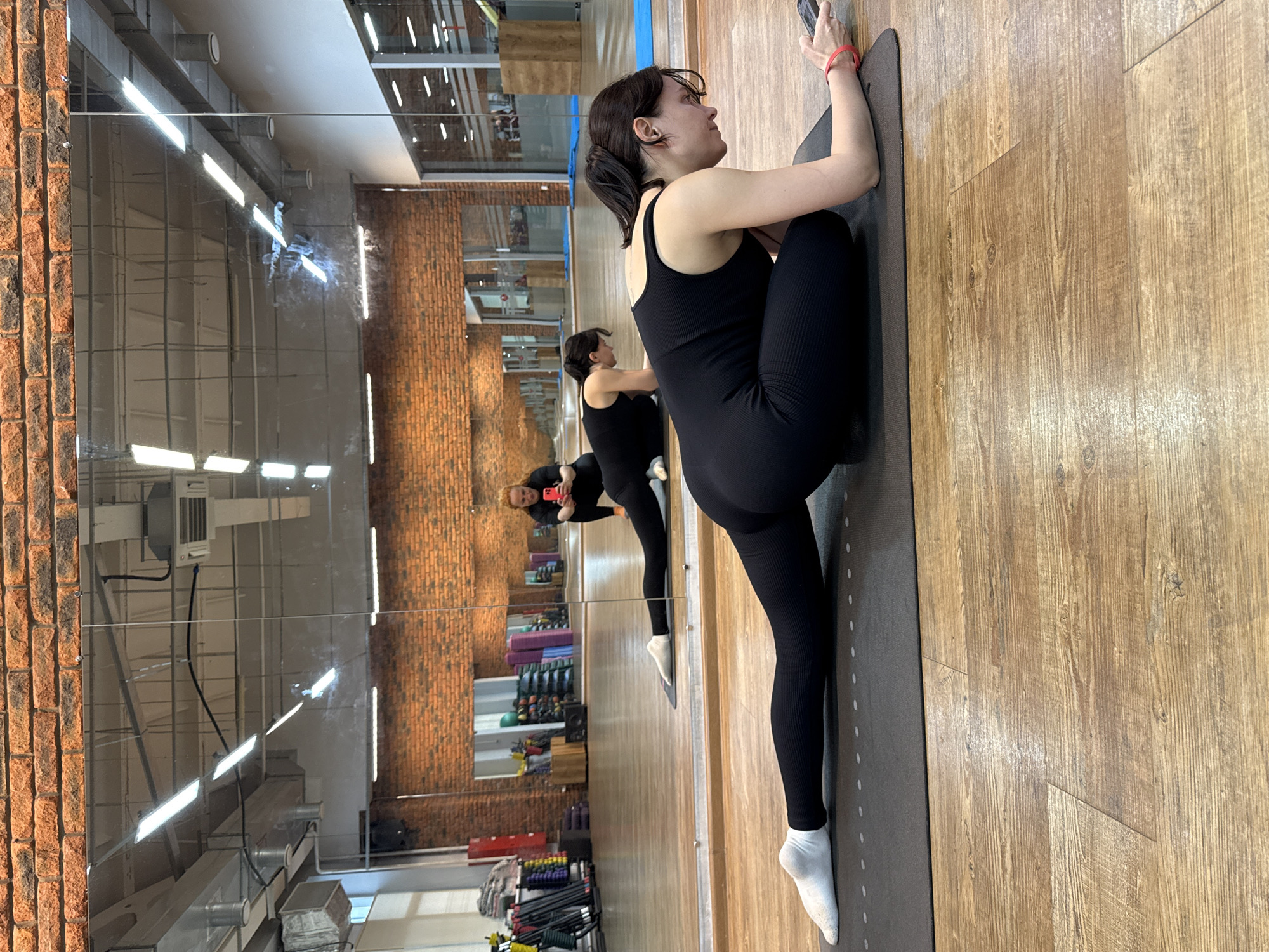 A woman in a black athletic outfit is performing a yoga pose on a mat in a studio. She is positioned on her right side, with her left leg extended upwards and bent at the knee, her left foot resting on her right shoulder. Her right leg is bent and tucked underneath her body. She is holding a smartphone in her right hand, and her left hand is resting on her left leg. Her expression is focused.
The setting appears to be a gym or fitness studio, with a mirrored wall reflecting the scene. The floor is made of wood. In the reflection, another woman in a similar black outfit is visible, also on a yoga mat, facing away from the camera and looking at her phone. Beyond the mirrored wall, shelves filled with exercise equipment like yoga mats, resistance bands, and weights are visible. The room is illuminated by overhead lighting. The overall impression is one of a personal fitness session or class.