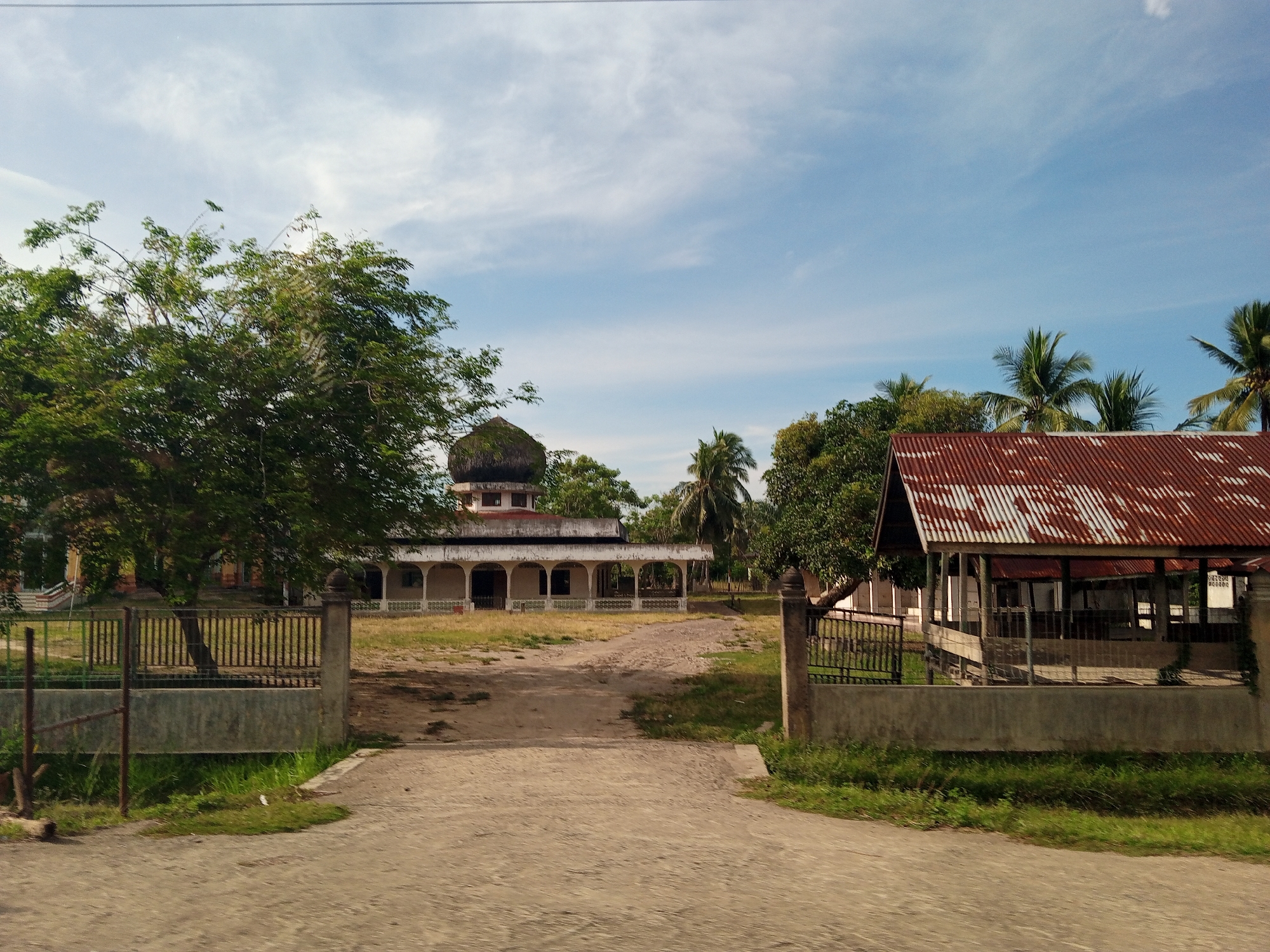 This image depicts an outdoor scene in Tumpokbeurandang, Indonesia, under a partly cloudy blue sky. The foreground features a light-colored, unpaved road or path leading into an open area. Flanking this entrance are two low concrete walls topped with metal fences; the left fence has green horizontal bars and brown vertical bars, while the right fence is darker, likely black or dark brown. Behind the fences and along the path, patches of green grass are visible, suggesting a somewhat unkempt environment.
In the midground, a wide, unpaved expanse of dry earth and sparse grass leads to two distinct structures. To the left, a light-colored building, possibly white or cream, stands with a prominent rounded, dark, possibly thatched or shingled dome on its roof. The building features an open veranda or porch supported by multiple arches. To the right, another building with a rusted, corrugated metal roof, predominantly reddish-brown with white weathered patches, is partially visible. This structure appears more open-sided.
The scene is heavily vegetated with various trees. A large, dense green tree is prominent on the left, partially obscuring the building behind it. Numerous palm trees and other green trees are scattered across the midground and background, particularly visible beyond both buildings, indicating a tropical or subtropical setting. No individuals or animals are present in the visible frame. The overall impression is a quiet, rural or semi-rural landscape.