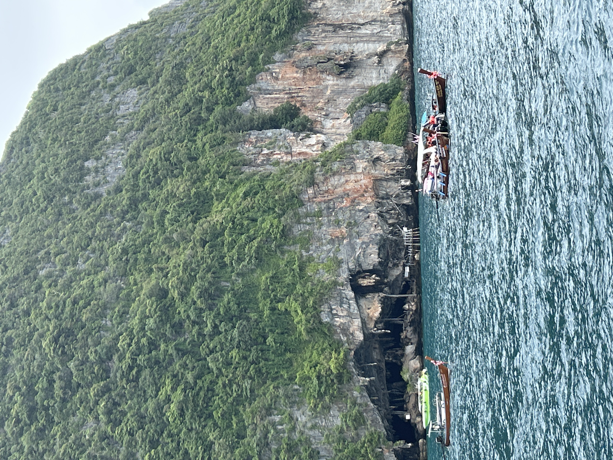 The image depicts a scenic view of a towering, lush green limestone cliff face with a large cave opening visible at its base, overlooking a turquoise body of water. Several traditional long-tail boats are anchored in the water, some carrying passengers. The boats are made of wood and feature the characteristic long outriggers. The cliff is densely covered in vegetation, with rocky outcrops and some visible striations in the rock. The water is choppy, indicating a gentle breeze. In the distance, a few more structures or boats can be vaguely discerned near the cave. The scene is likely a tourist destination in the Ao Nang area of Thailand, possibly a well-known cave or island attraction.