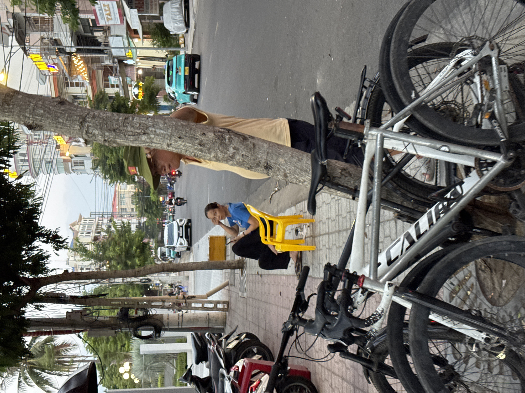 This image depicts a street scene in Nha Trang, Vietnam, with a focus on the sidewalk and the adjacent road. In the foreground, two bicycles are parked, one silver and white, the other darker. Leaning against a tree on the sidewalk, a man wearing a green military-style cap and a yellow shirt stands with his back mostly to the camera. To his left, a woman in a blue shirt and dark pants sits on a bright yellow plastic chair, looking down at her phone.
The sidewalk is paved with concrete tiles and lined with a curb. Various power lines and cables are strung across poles along the street. Parked motorcycles are visible on the sidewalk further down. The road features vehicular traffic, including a white car, a turquoise car, and several motorcycles. Buildings line the opposite side of the street, displaying signage and architectural details typical of a commercial area in Vietnam. The overall atmosphere suggests a typical day on a busy urban street.