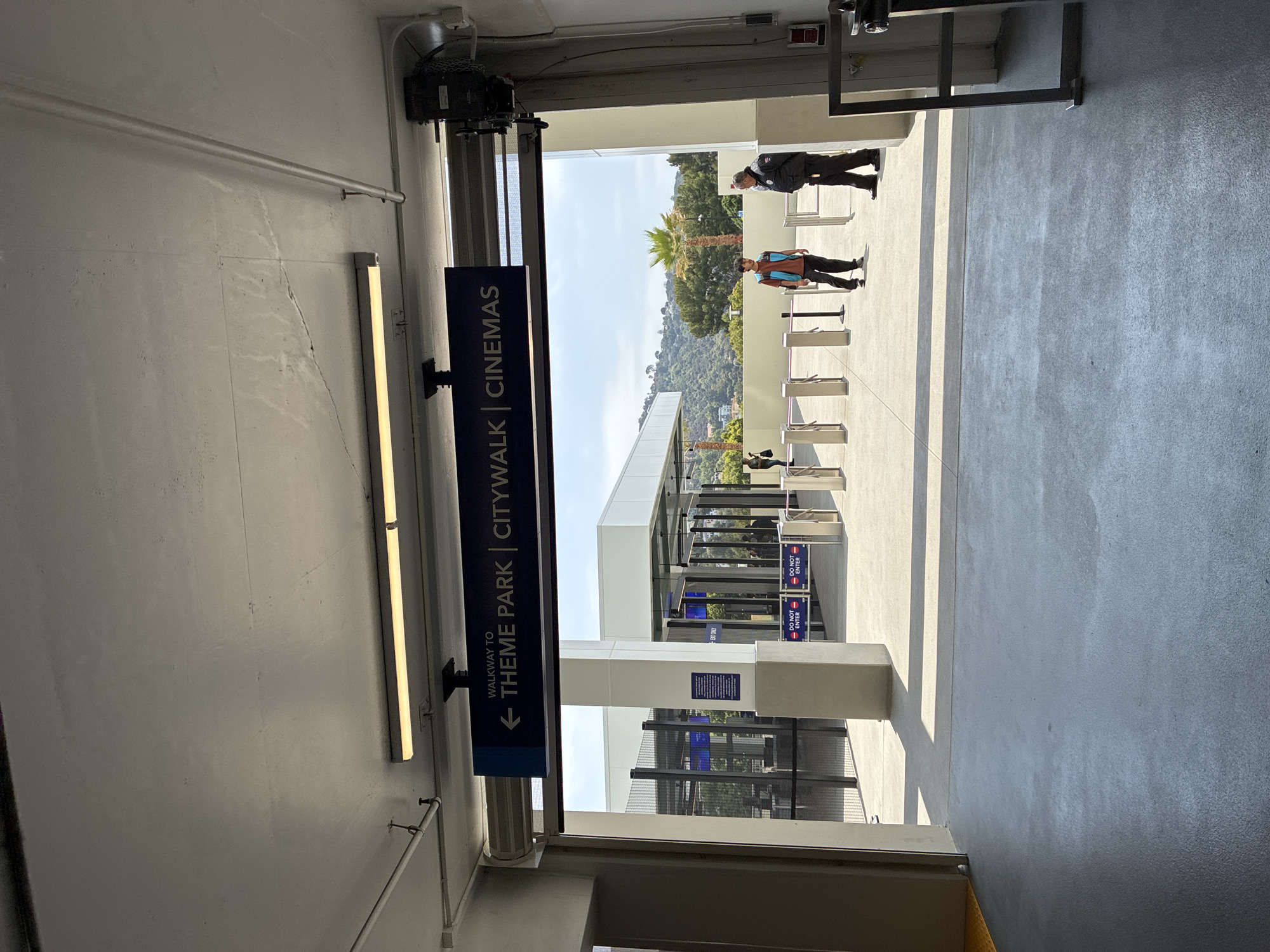 An exterior, eye-level, medium shot shows an entrance to a pedestrian walkway that leads to a theme park and cinemas. The walkway is covered by a concrete overhang with fluorescent lighting. A dark blue sign with white text hangs from the overhang, reading "WALKWAY TO ← THEME PARK | CITYWALK | CINEMAS."
Beyond the entrance, the walkway opens to a sunlit area with multiple ticket turnstiles. Two men are visible near the turnstiles. One, wearing a dark jacket and pants, appears to be a security guard. The other, a younger man in a teal vest over an orange shirt and black pants, stands facing the security guard. Behind the turnstiles, a tall fence encloses an area with more structures and trees. In the background, a lush green hillside rises under a clear blue sky with scattered clouds. The environment suggests a warm, sunny day in a location with a mild climate. The overall scene depicts an access point to a public entertainment venue.