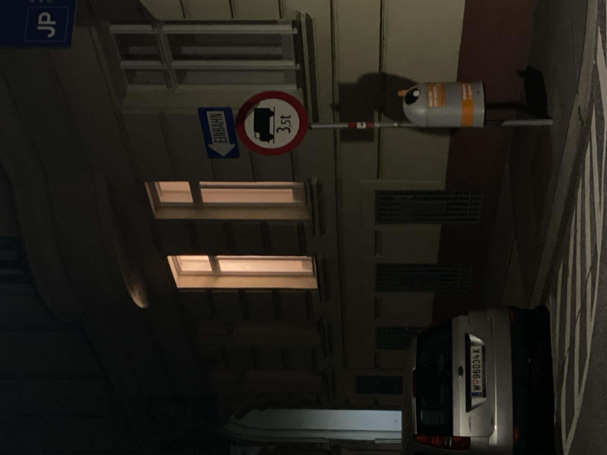 The image shows a street scene at night. A silver vehicle with license plate "W96034K" is parked at the right. Above the car, on the left side, are windows illuminated from the inside, and a parking permit with letters "JP". Above the windows is a traffic sign indicating "EINBAHN" (one way) with an arrow pointing down, and to its right is a weight limit sign of "3,5t". To the right is street lighting. The location appears to be an urban area.