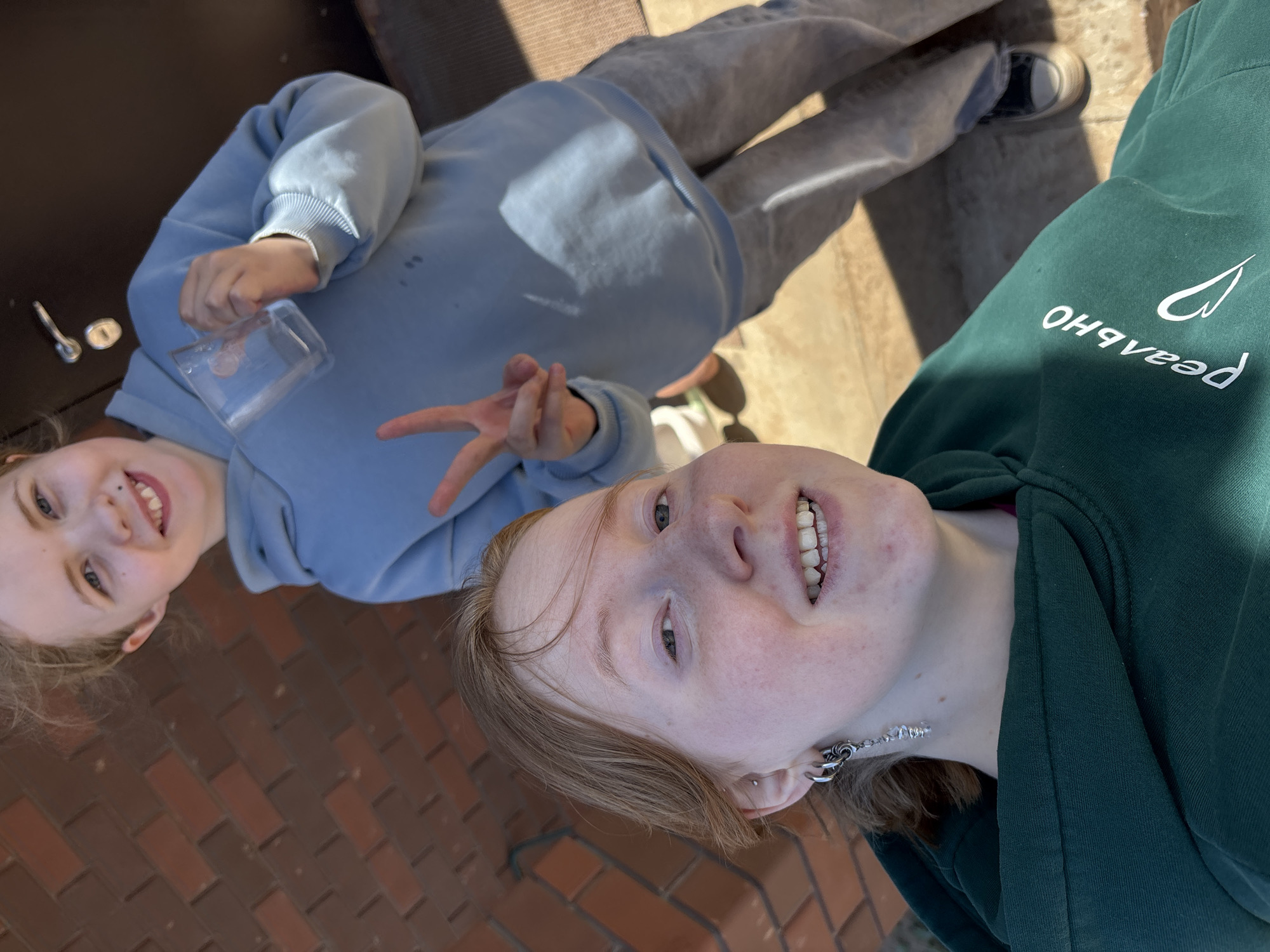 Two individuals are depicted outdoors on a brick surface in Prikamskii, Russia. One individual wears a light blue hooded sweatshirt and light grey sweatpants with dark shoes. This person holds a clear glass container. The second individual wears a dark green hooded sweatshirt and distinctive earrings. The hoodie's front has text mirrored and vertically arranged. They are positioned with the brick surface directly beneath them, with the background comprising concrete or tile surfaces. The lighting is bright, indicating outdoor daylight.