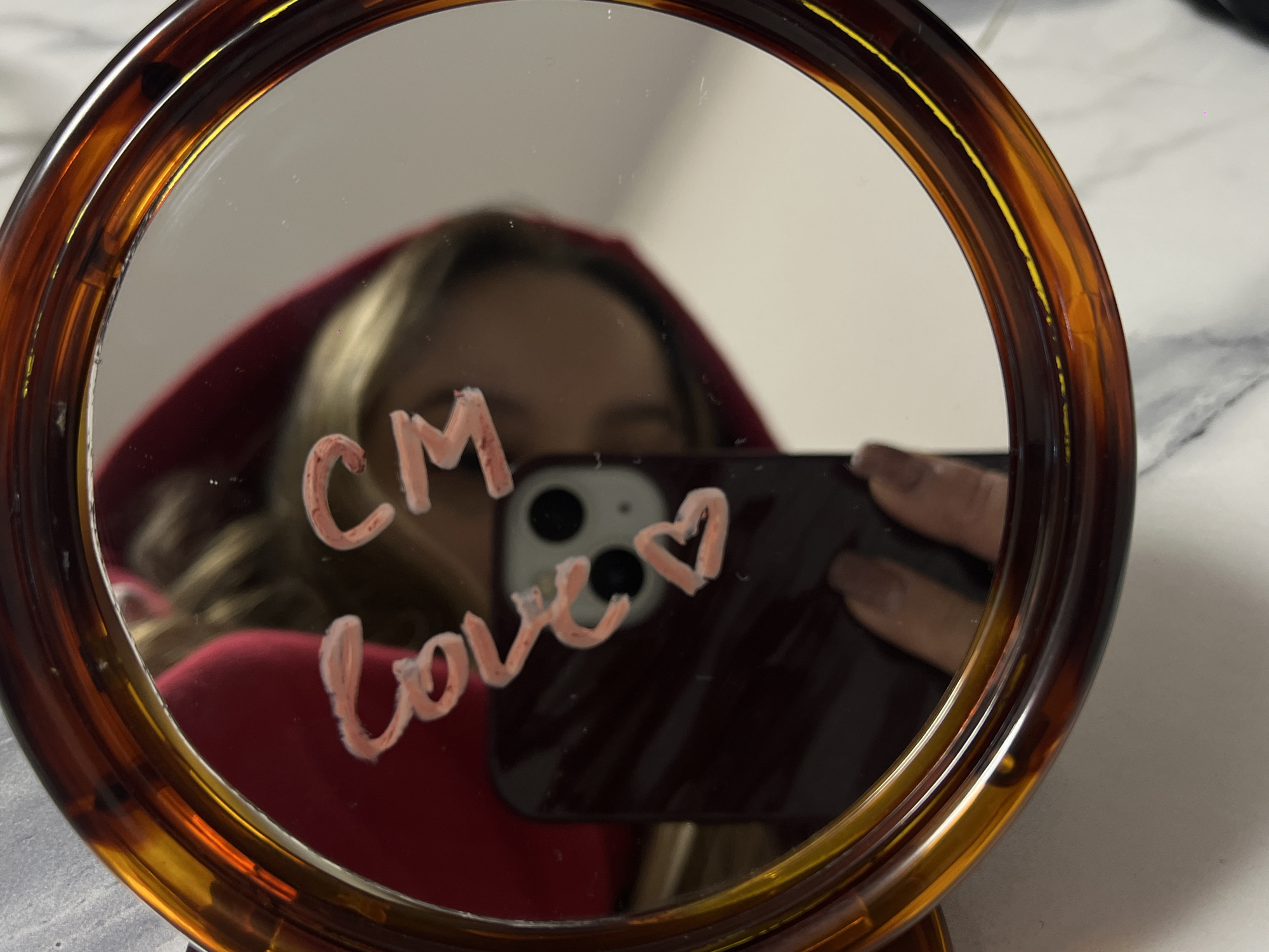 A close-up, eye-level shot captures a person's reflection in a round mirror with a brown and amber tortoiseshell frame. The mirror serves as the central subject, reflecting a person holding a smartphone, presumably taking the picture. The person's face is obscured by the phone, but their blonde hair and a red hooded garment are visible. The smartphone, visible as a dark rectangle with two prominent circular camera lenses on its back, is held up to capture the reflection.
Written on the mirror's surface in a light pink or peach color is the text "CM love ♡", with a drawn heart symbol. The reflection outside the mirror's frame shows a light-colored, possibly marble-patterned surface, while the background reflected within the mirror appears to be a plain light wall. The image provides an intimate glimpse of a moment of self-expression or affection, set indoors in Krasnodar, Russia. The focus is sharp on the mirror's surface and the writing, with the reflected person appearing slightly softer.