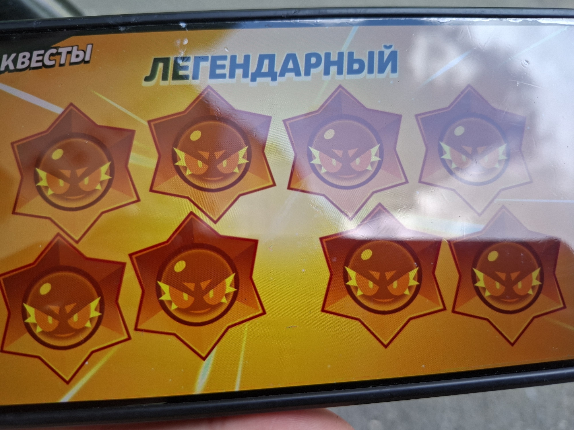 A close-up photograph captures a screen or printed material displaying game-related graphics. The dominant colors are vibrant oranges, yellows, and reds. Six star-shaped badges are prominently featured, arranged in two rows. Each badge has a bright orange outline and a darker red interior, containing a circular design with a stylized, angry-looking face. This face consists of two glowing orange eyes, a small nose, a downturned mouth, and jagged yellow elements on either side, possibly representing sparks or anger.
The background is a gradient of orange and yellow, with subtle white diagonal lines or streaks. Visible text at the top is in Russian: "КВЕСТЫ" (QUESTS) is partially visible on the left, and "ЛЕГЕНДАРНЫЙ" (LEGENDARY) is clearly displayed in blue letters to the right.
Significant glare and reflections are present across the surface, particularly noticeable in the upper right portion, obscuring some details of the badges and background. The image is framed by a black border, suggesting it's a photo of a device screen, with parts of fingers visible at the bottom edge. The overall aesthetic suggests content from a mobile game, possibly Supercell's Brawl Stars, given the style of the star icons.