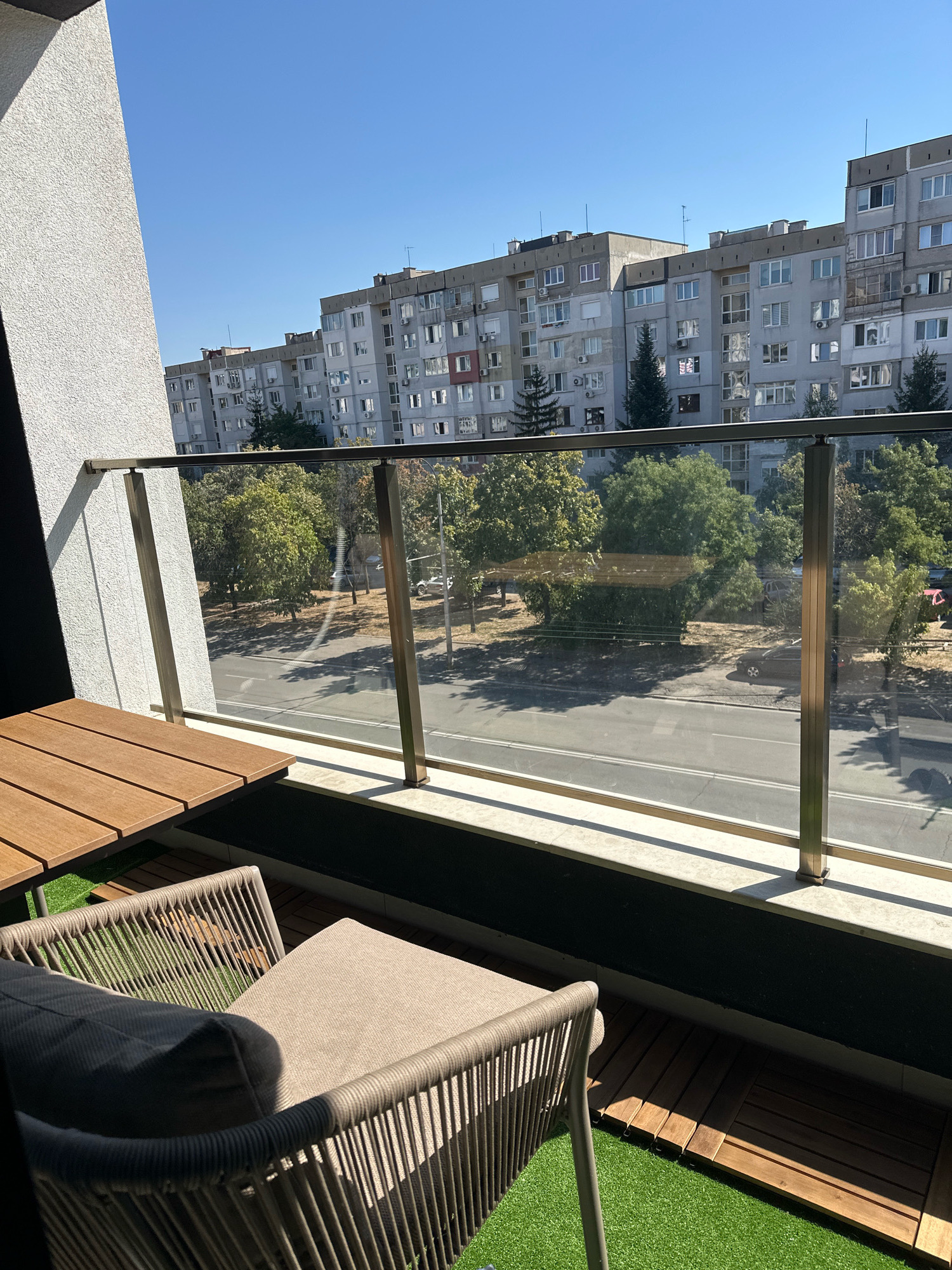 The image shows a view from a balcony in Sofia, Bulgaria, overlooking a street with apartment buildings and trees. In the foreground, there is a modern balcony set with a wooden table and a cushioned chair with a gray pillow. The balcony floor is covered with artificial grass and wooden decking. Beyond the glass railing, a street is visible with traffic and parked cars. Several multi-story residential buildings, characteristic of Soviet-era architecture, line the street, with trees and greenery interspersed between them. The sky is clear and blue, indicating a sunny day.