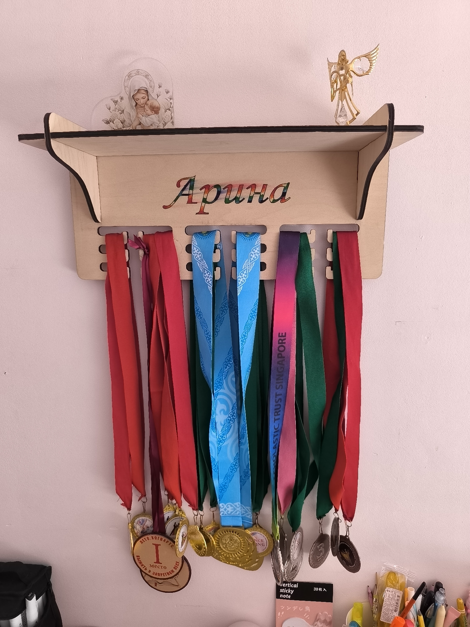 The image shows a wooden shelf mounted on a pink wall, used to display a collection of medals. The shelf is decorated with a small, heart-shaped image of the Virgin Mary and a golden angel figurine. Below the shelf, ten medals hang from hooks, each attached to a colored ribbon. The medals vary in size, material, and design, with some featuring inscriptions in Russian and English. To the left of the shelf, a black bag is partially visible. To the right, a collection of stationery items, including pens, pencils, and sticky notes, are arranged on a surface. The overall scene appears to be a personal display area, possibly in a home, in the city of Oskemen, Kazakhstan.