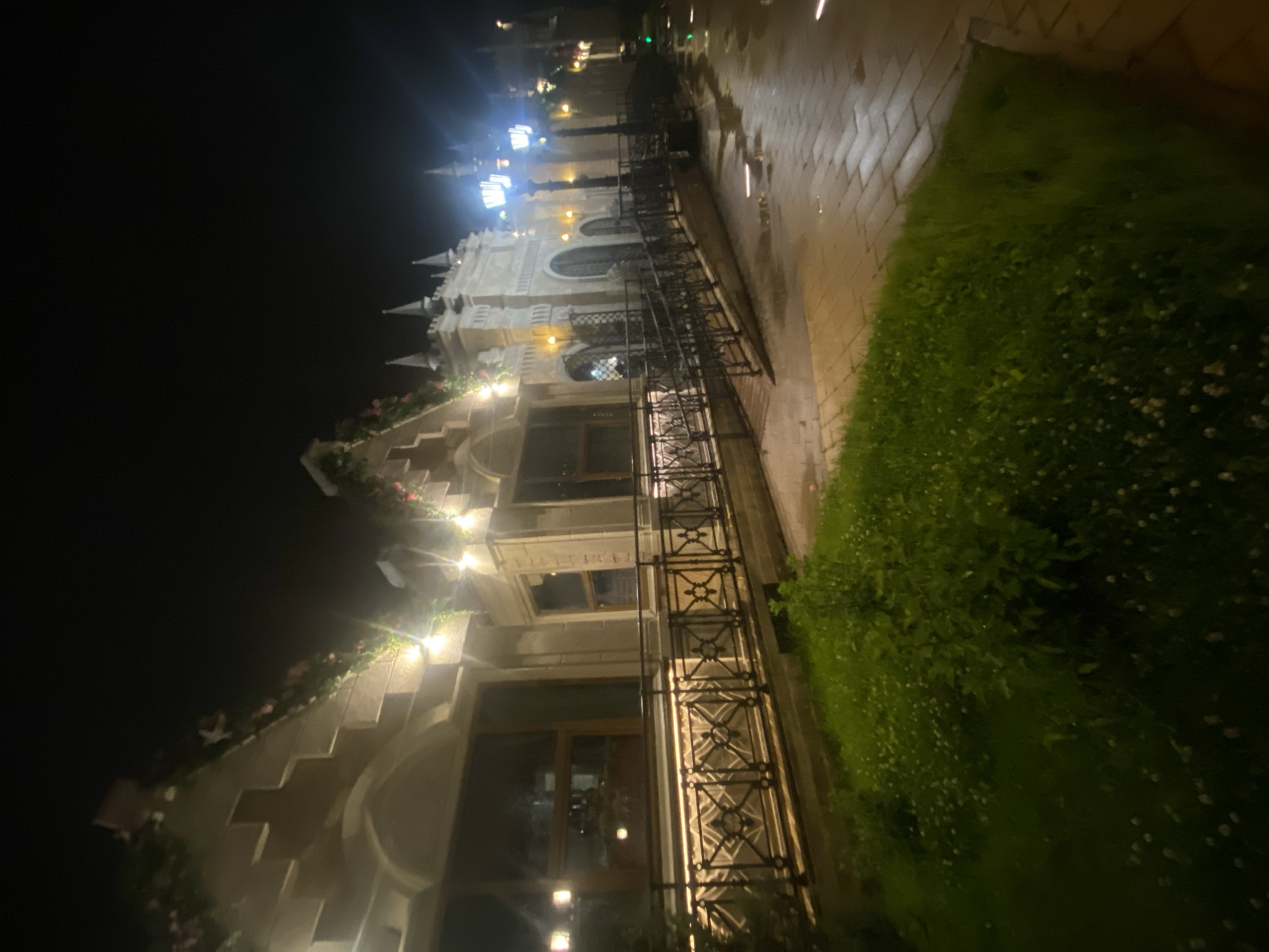 The image shows a building with ornate architecture and decorative lighting at night, suggesting a restaurant or entertainment venue in Moscow, Russia. The building features pointed roofs, decorative stonework, and large windows. String lights are visible along the facade and on the rooftop, casting a warm glow. In the foreground, there is a well-maintained grassy area with small white flowers. A paved walkway with decorative streetlights runs alongside the building. The ground appears wet, indicating recent rain. There are no people visible in the image.