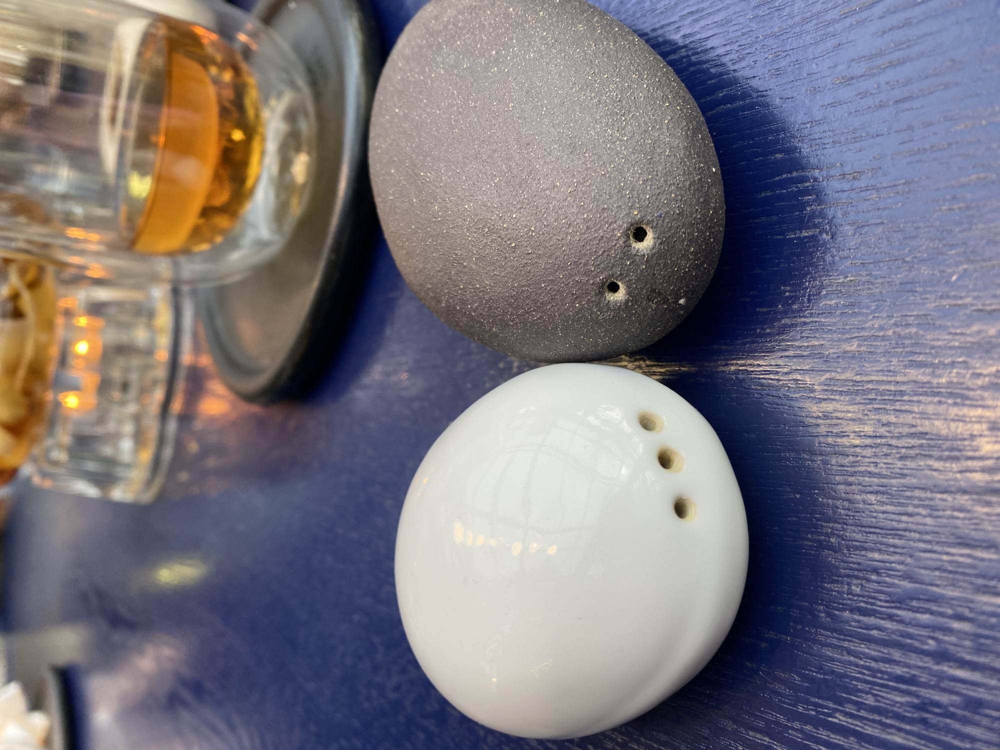 The image shows a close-up of a dining table setting, likely indoors. A pair of salt and pepper shakers is prominently featured, one gray with two holes and the other white with three holes. To the left are partially visible drinking glasses with a yellowish liquid, potentially alcohol, sitting on top of a black plate. The entire scene is set against a blue, textured, possibly wooden table surface. The specific location in Yekaterinburg, Russia cannot be determined from the provided visual information. There are no visible people or interactions in the scene.