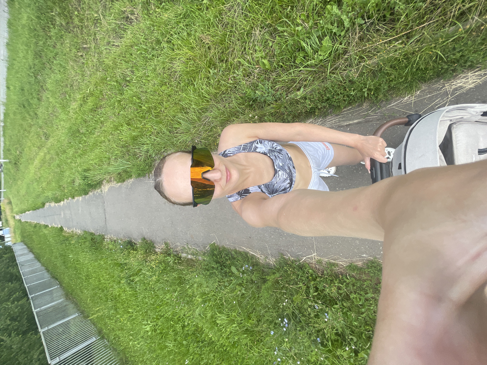 A woman wearing a sports bra, grey shorts, and reflective orange-lensed sports sunglasses is pushing a stroller along a narrow paved path. The path is bordered by lush green grass on both sides. To the left of the path is a metal fence. The woman is holding the stroller handle with her right hand. The perspective is a first-person point of view, as if the viewer is the woman. The setting appears to be a park or a suburban area on a cloudy day. The image is taken in or near the city of Podshivalovo, Russia.