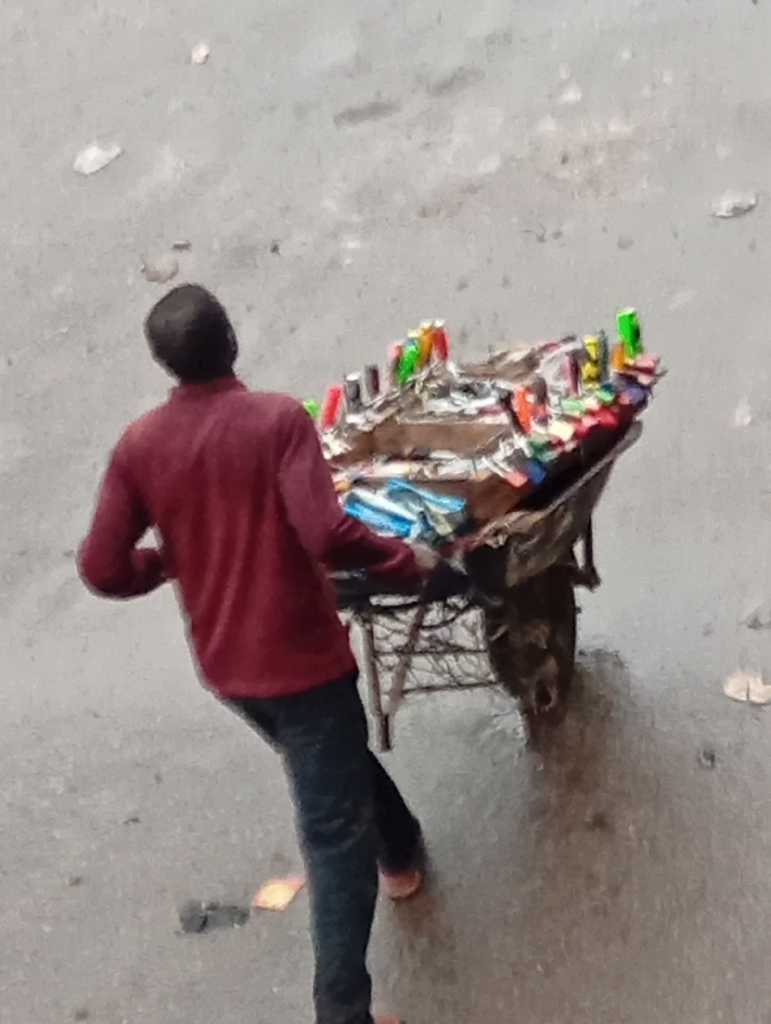 A man in a red shirt and dark pants is standing next to a cart laden with various colorful items, possibly for sale. The scene is set on a paved surface, likely a street in Bukuru, Nigeria. The cart appears to be homemade or simple in construction. The man is facing towards the cart. The overall impression is that of a street vendor in a marketplace setting.