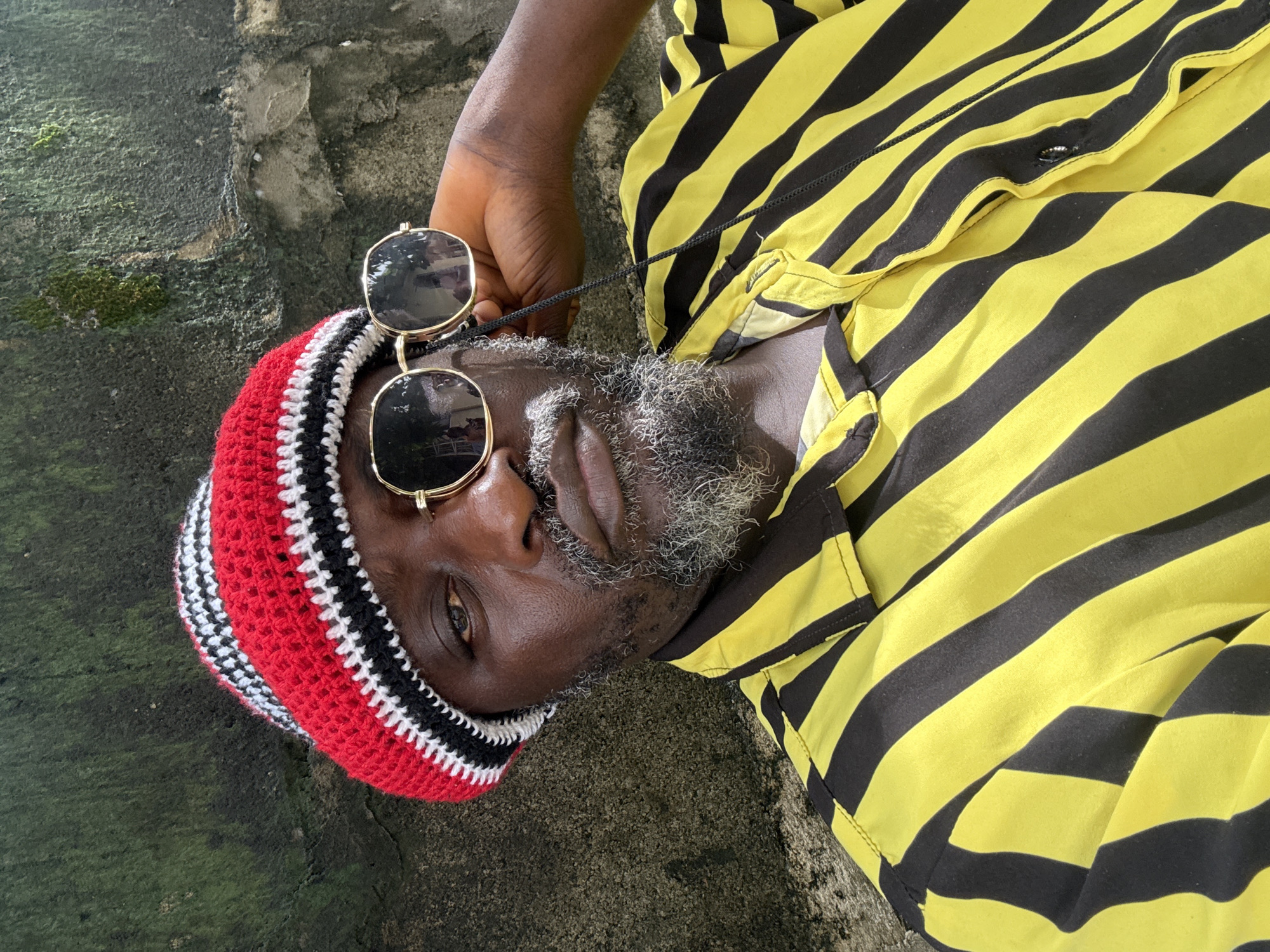 An eye-level shot from a slight angle, shows a man with graying facial hair lying on a concrete surface. He is wearing a red, black, and white crocheted hat and sunglasses with gold frames. His shirt has yellow and black horizontal stripes. He is lying on a concrete surface that appears to be part of a weathered wall or floor, possibly in Ado, Nigeria.