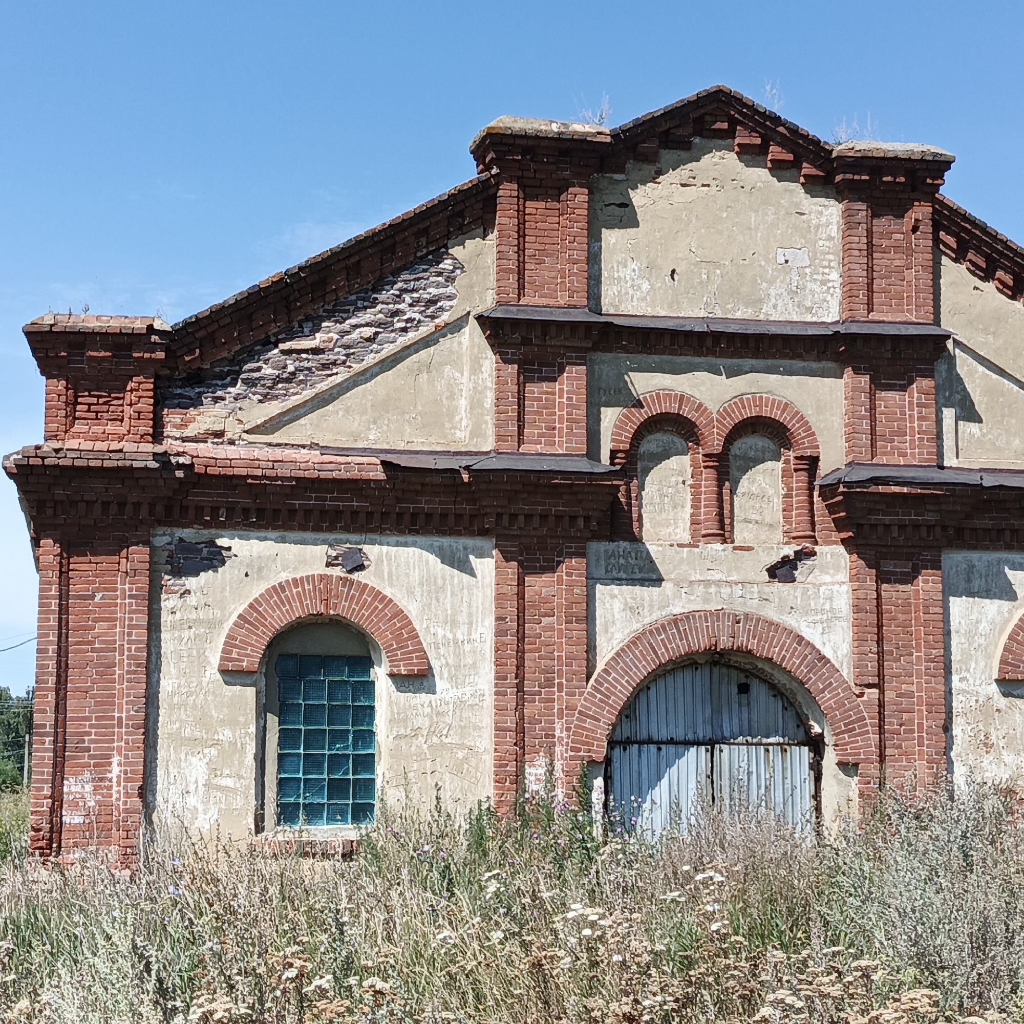 The image shows the exterior of a weathered, abandoned brick building in Тугустемир, Russia. The building has a damaged roof, with exposed brickwork and crumbling plaster. The facade features arched windows and doorways, some with decorative brickwork. One window has a grid of blue glass blocks. The lower part of the building is obscured by overgrown, dry vegetation and wildflowers. The sky is clear and blue. There are no people visible in the image. The overall impression is one of decay and neglect.