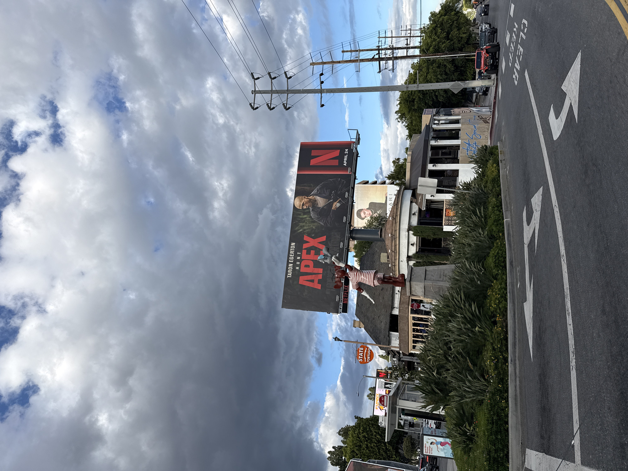 The image is taken in West Hollywood, United States of America, facing an Apex billboard. The billboard features Taron Egerton and the words "APEX" and "HUNT," plus the date "APRIL 24" and the Netflix logo. Next to the Apex billboard is another billboard for Bulgari. Below the billboards, there is a building with retail establishments, including a State Tortugo restaurant and shop. A figure of a man in a striped shirt is on top of one of the buildings. The street is asphalt, with a lane marked with a large white arrow indicating the direction of traffic. Cars are parked along the street. A row of bushes and foliage separates the street from the buildings. Above the billboards are power lines, and the sky is filled with dark, partly cloudy.