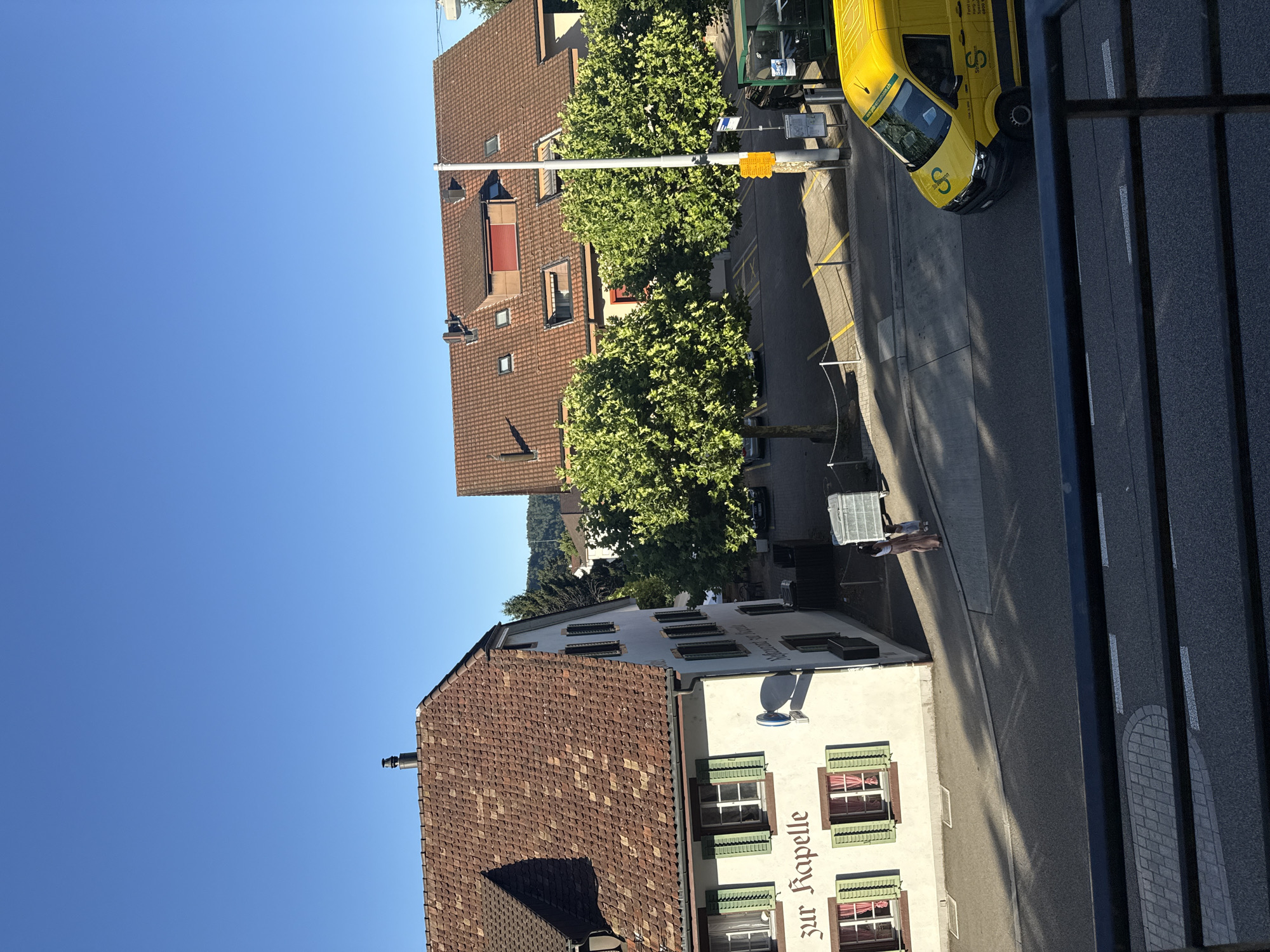 The image captures a street scene in Trimbach, Switzerland, on a clear day. In the foreground, a black metal railing overlooks a road. To the left, a light-colored building with a red-tiled roof features green shutters on its windows and signage that reads "zur Kapelle." To the right of this building, a parking lot is visible with several trees providing shade. Further to the right, another building with a red-tiled roof is present, characterized by dormer windows and a red shutter. A street light stands in front of this building. A yellow van with a logo that includes the letter "S" is driving on the road. In the mid-ground, two individuals are standing on a sidewalk near the first building. The environment is urban with residential and commercial buildings, trees, and a road.