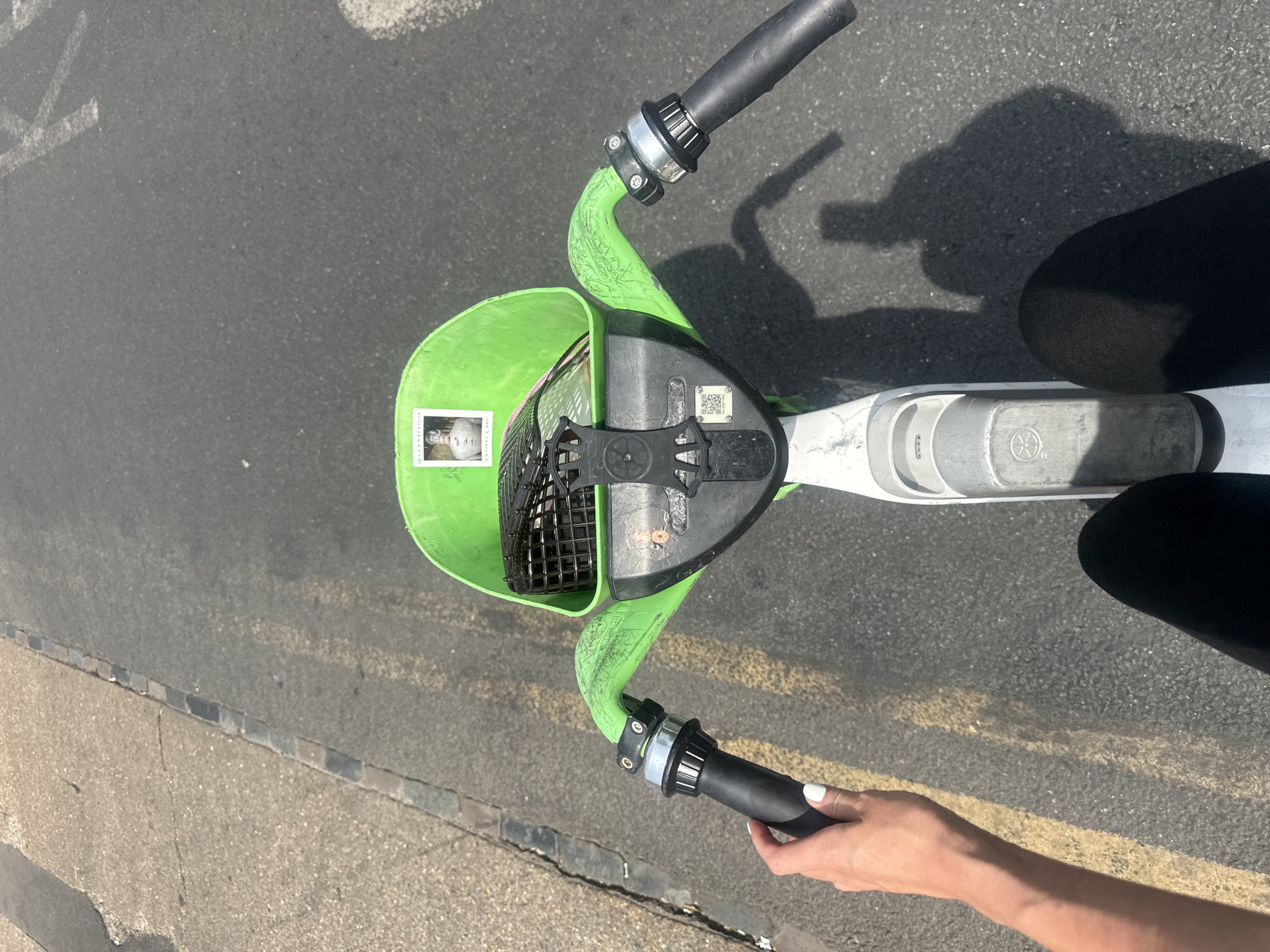 The image shows the handlebars of a Lime scooter on a paved surface. The scooter is light green with black handlebars and a black basket with a black mesh interior. There is a rectangular sticker on the front of the basket and a QR code near the handlebar attachment. The scooter is being held by a light-skinned hand with white nail polish. There is a shadow of the scooter rider visible on the pavement. The pavement has yellow lines painted on it. Based on the scooter type and the overall look, this is potentially taken in London, UK, as Lime is a dockless scooter rental company that operates in many cities, including London. However, without additional context, a definitive location confirmation is not possible.