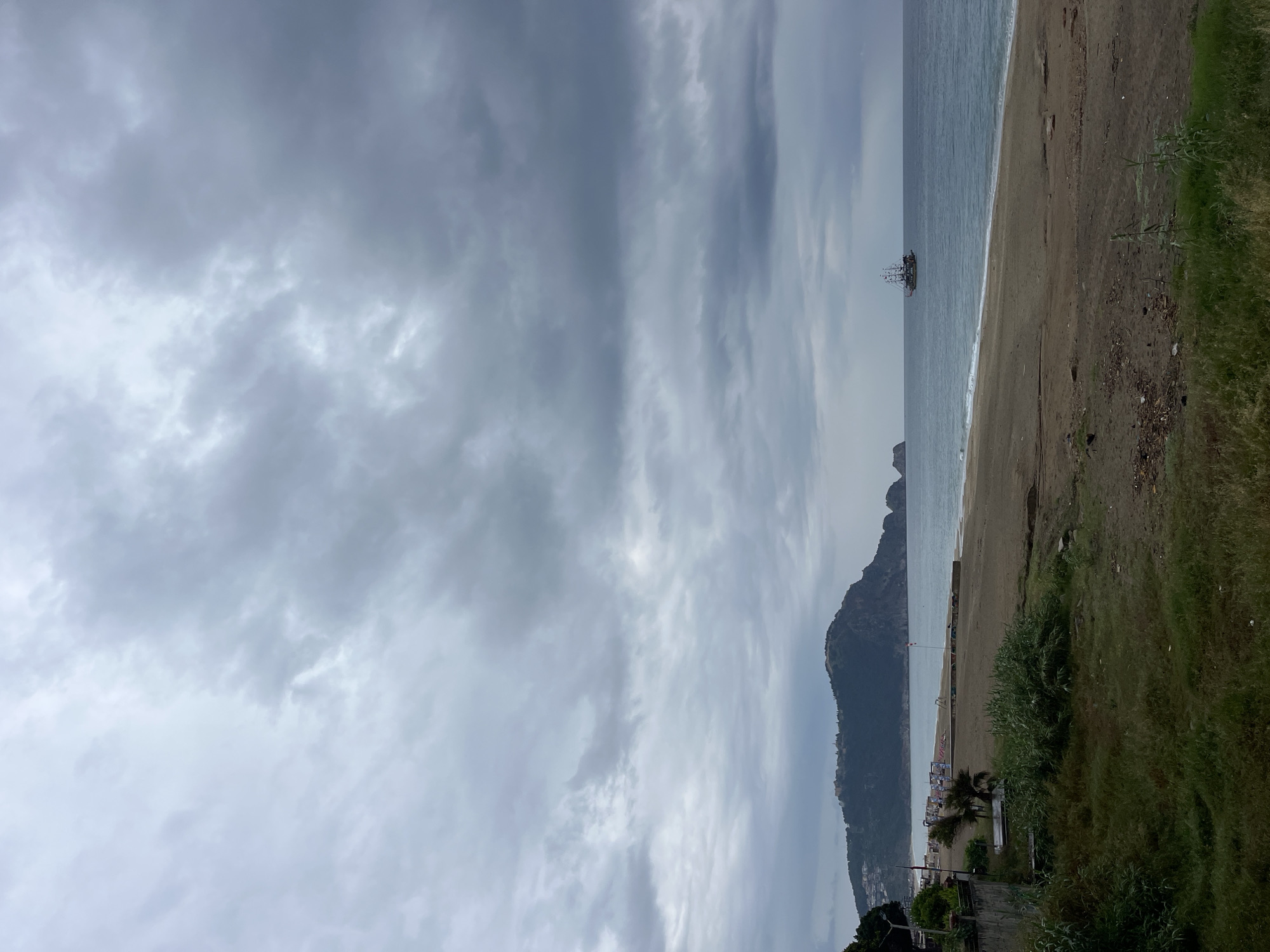 The image captures a coastal scene in Alanya, Turkey, under a cloudy sky. A sandy beach stretches along the coastline, with gentle waves meeting the shore. A mountain rises in the background, partially obscuring the view of the city. A ship is visible on the water. Vegetation grows along the edge of the beach.