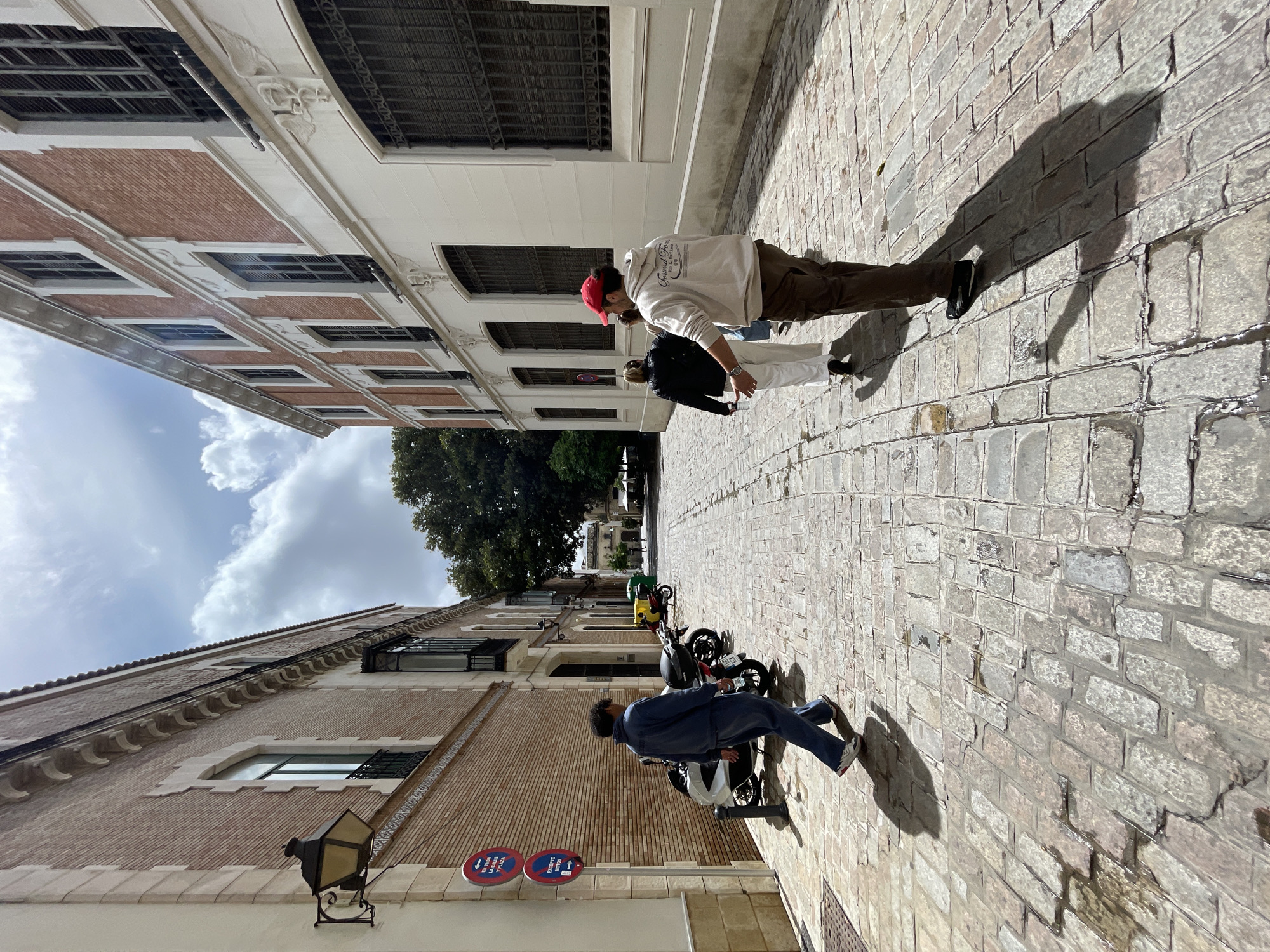 A sunny day captures a narrow street in Jerez De La Frontera, Spain, framed by buildings on either side. The street is paved with cobblestones. On the right side of the image, two people are walking away from the camera. The first person is wearing a red cap, a white hoodie, and brown pants. The second person has black clothing and white pants. On the left, a man wearing a blue outfit sits atop a scooter. Further down the street, two motorcycles are parked near green trash cans. The sky has scattered clouds. The buildings are constructed with brick and off-white stone accents. A lamppost is on the left building, along with a pair of signs displaying red circles and arrows. Shadows from the sun are cast across the cobblestone street.