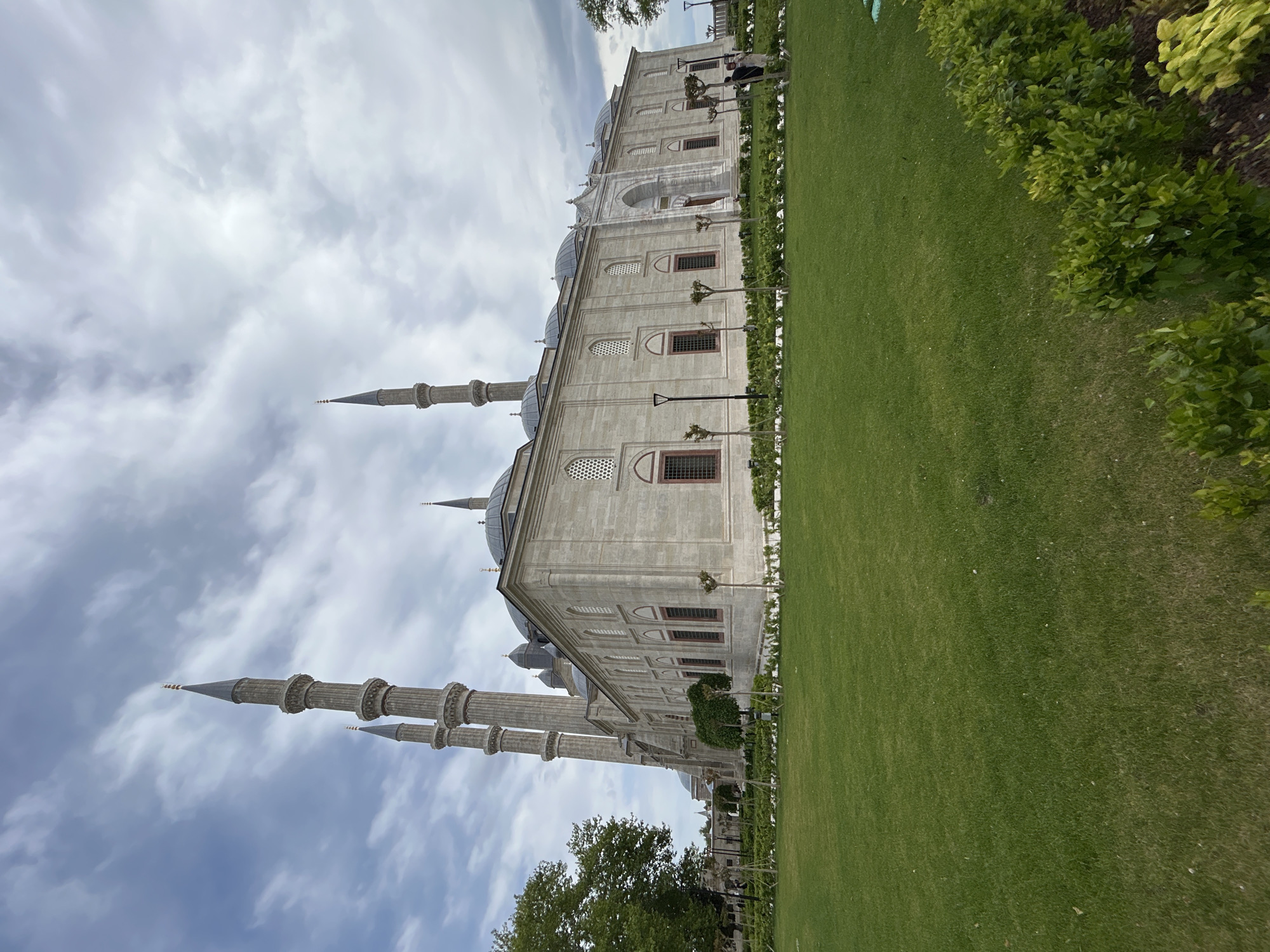 The image depicts the Selimiye Mosque in Edirne Merkez, Turkey. The mosque is a large, light-colored stone structure with multiple domes and tall, slender minarets reaching into the sky. The minarets are cylindrical with detailed, layered designs. The mosque is surrounded by a well-maintained green lawn and rows of bushes and small trees. The sky is filled with clouds, indicating either an overcast or partly cloudy day.