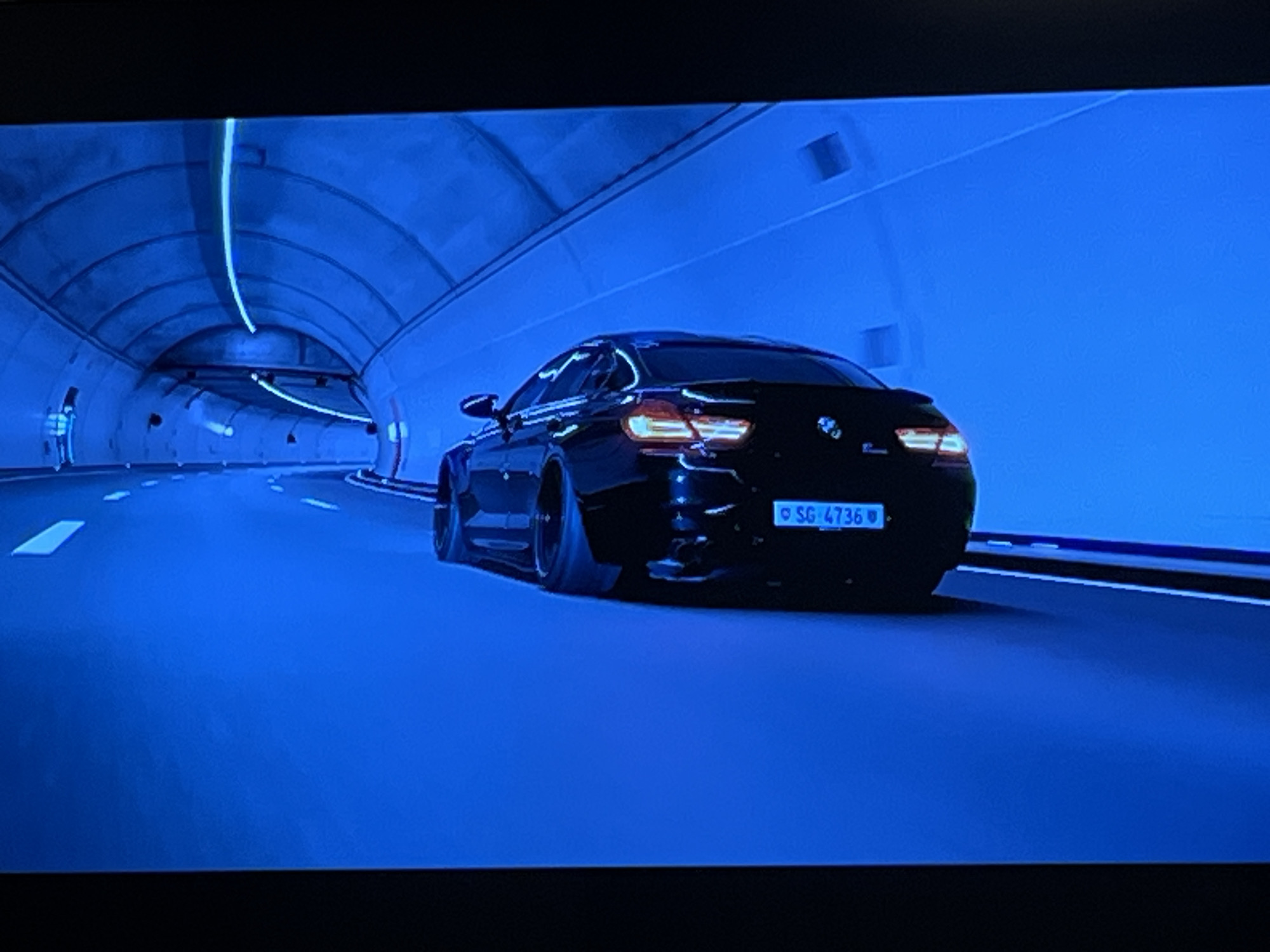 The media file displays a black car driving through a blue-lit tunnel. The car is seen from the rear, with its taillights illuminated. The tunnel has a rounded ceiling with integrated lighting that casts a cool blue glow on the walls and road. White dashed lines mark the lanes on the road, indicating it is a highway or a similar thoroughfare. The car appears to be moving at a considerable speed, suggested by the motion blur and the perspective of the tunnel.
The license plate of the car is visible and reads "SG 4736". The overall aesthetic is modern and somewhat futuristic, with the monochromatic blue lighting creating a dramatic atmosphere. There are no people visible in the image, only the car as the central object. The setting is clearly a tunnel, designed for vehicle traffic. The time of day and weather are not discernible as the scene is entirely within an artificial environment. There are no specific emotional cues conveyed by the image, apart from the potential for excitement or intensity associated with a car in a tunnel. The location context of Kharkiv, Ukraine, is not directly evident from the visual elements of the image itself, but is provided as external information.