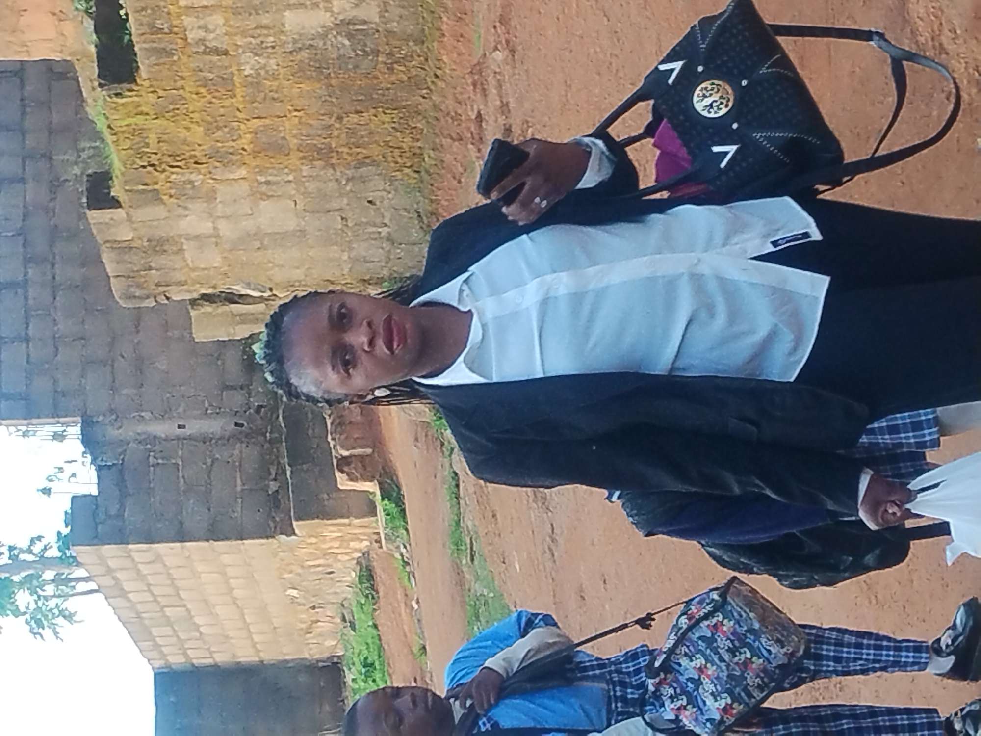 A medium shot shows a Black woman, likely in her late 20s or 30s, walking toward the viewer. She wears a dark blazer over a white button-down shirt and black pants. Her hair is styled in a braid, and she has large earrings. In her right hand, she holds a smartphone, and in her left, she carries a black handbag with a decorative emblem and shoulder strap. She is walking alongside two children. The child on the left, also Black, is wearing a blue shirt and a plaid skirt. The child on the right is partially obscured, but appears to be wearing a blue uniform. The scene is set outdoors on a dirt path, with a stone wall and a building in the background. The environment suggests a rural or semi-urban area.